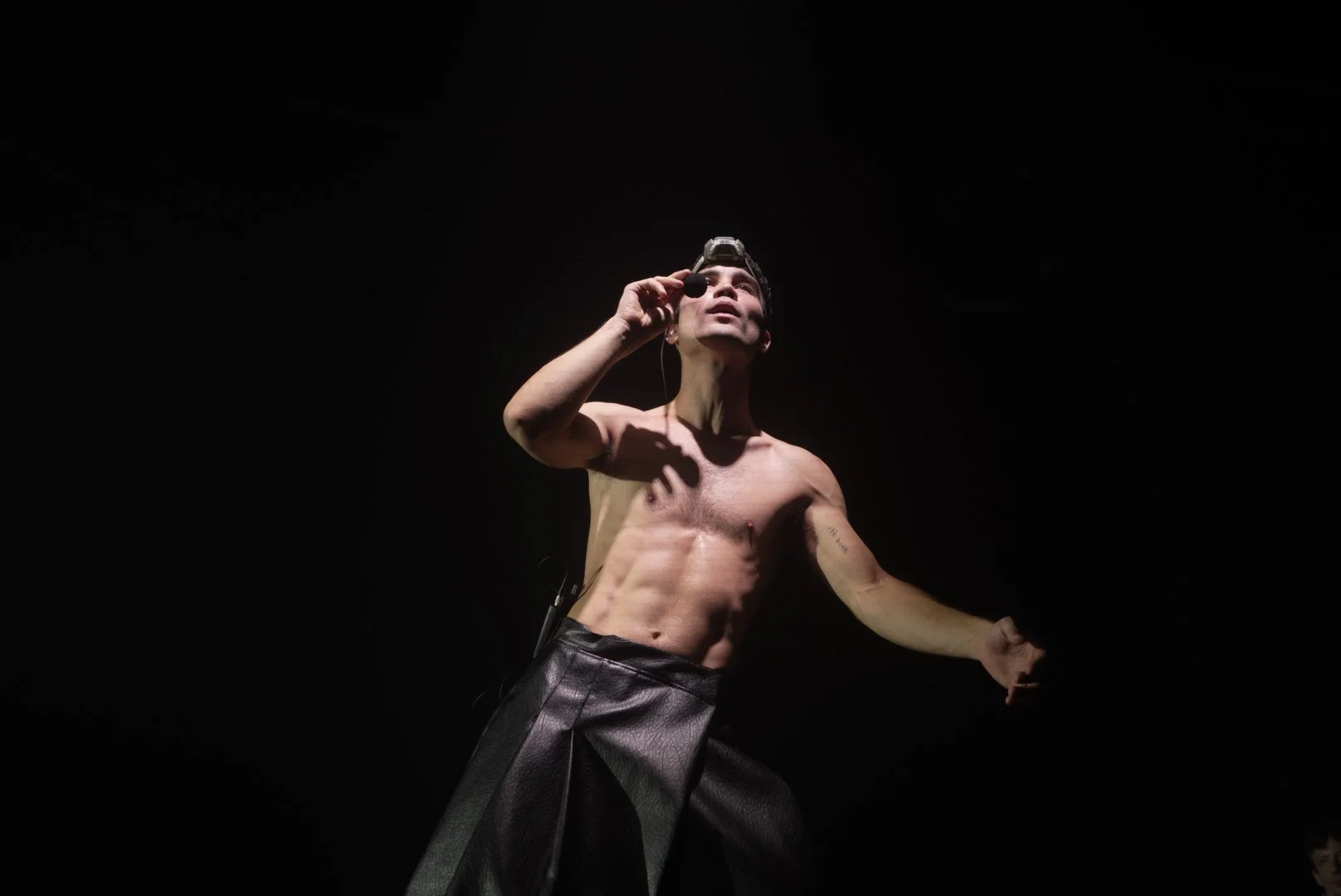 A shirtless male performer on stage, holding a microphone close to his mouth, wearing a headlamp and black leather pants, with a dark background.