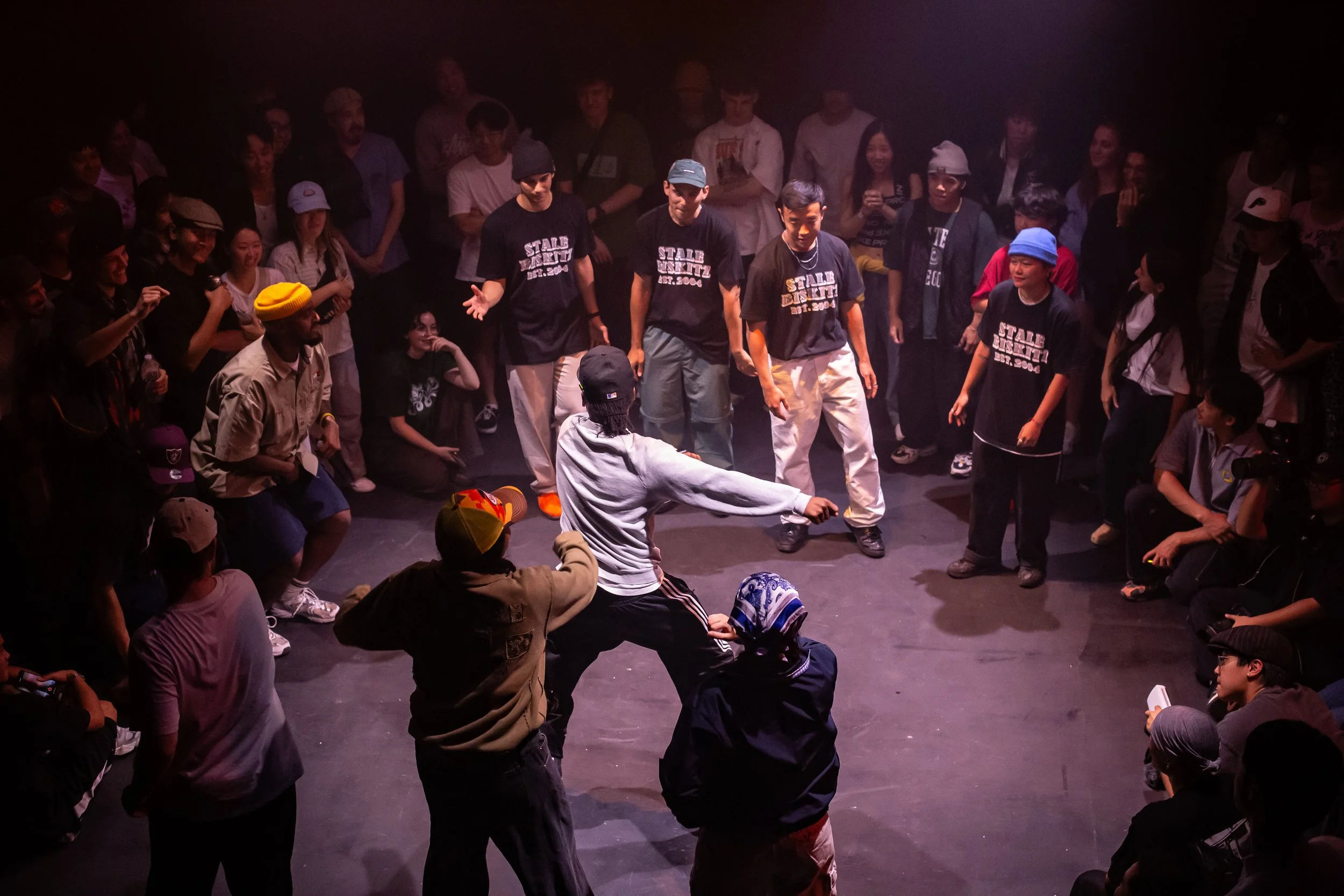 A dance battle in a circle with people surrounding and watching, some taking photos, in a dark indoor setting with spotlights.