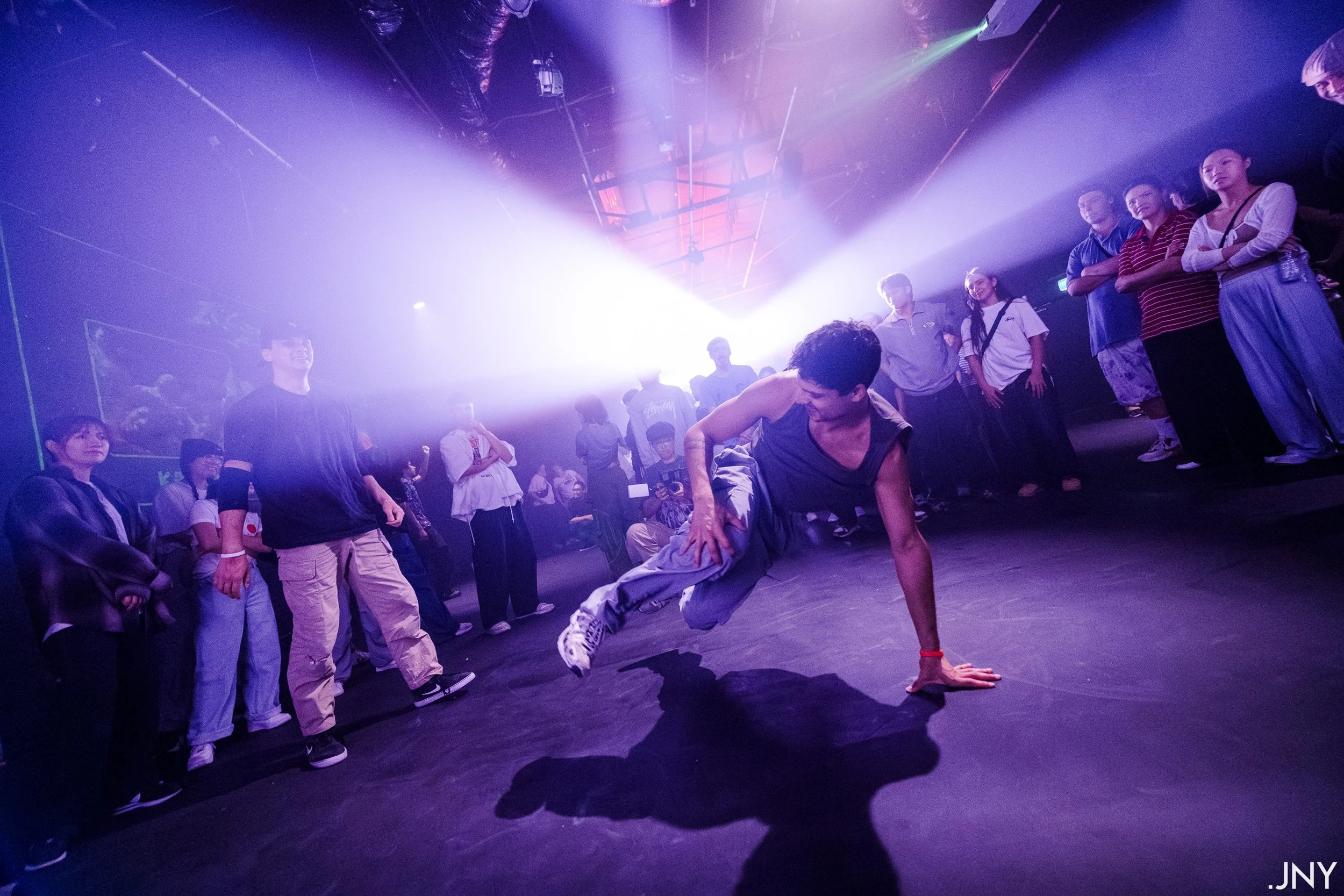 A young man performing a breakdance move on a stage surrounded by an audience, with purple and white stage lighting creating a vibrant atmosphere.