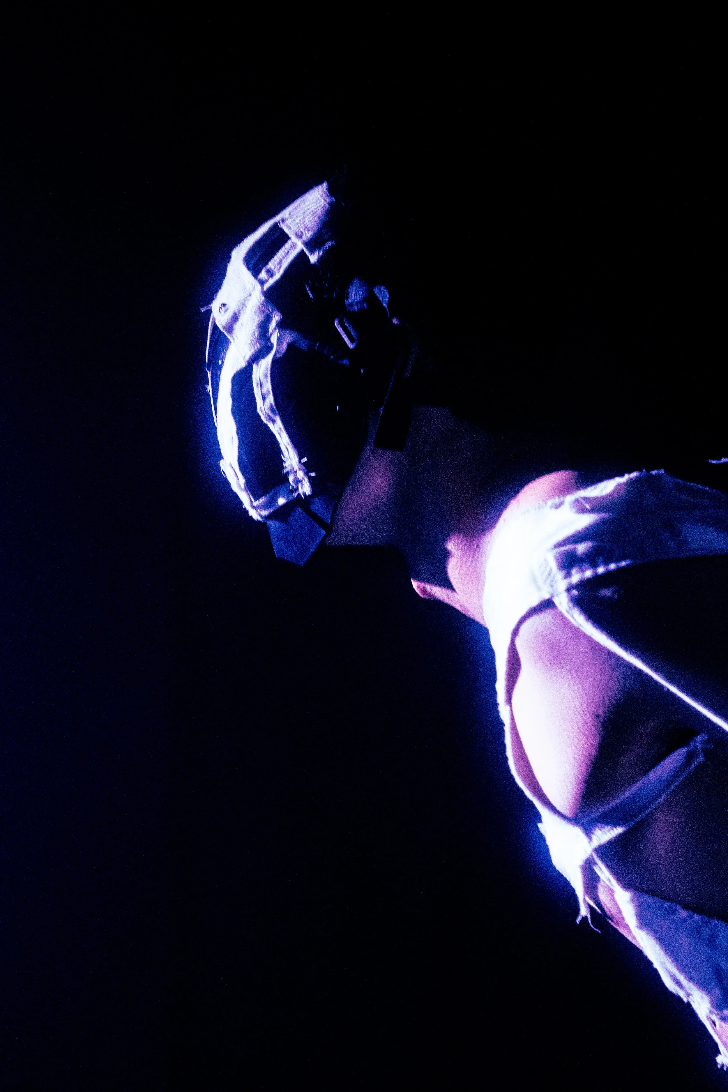 A person’s back and side profile, wearing a black cap, denim shorts, and a white shirt, illuminated by blue and purple stage lighting.