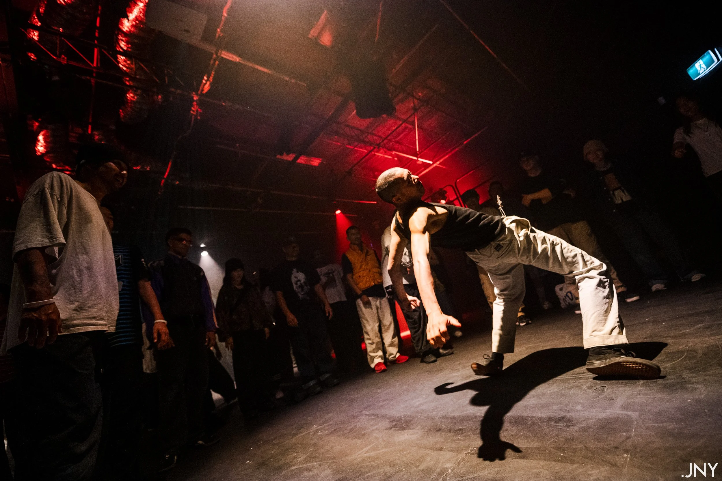 A man performing a breakdance move on a dark stage with red lighting. Several people watch him.