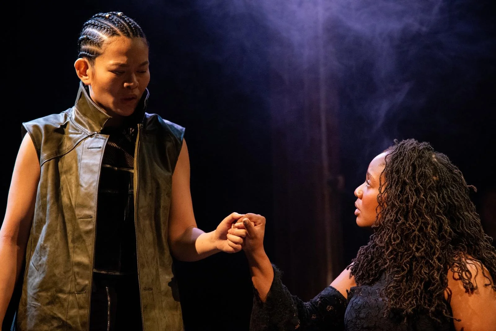 Two women exchanging a handshake, with one woman standing and the other sitting, in a dimly lit setting.