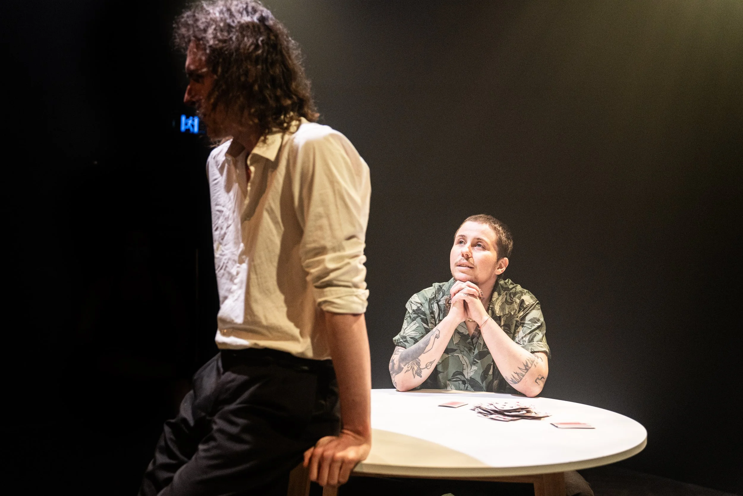 Two men are involved in a scene with one sitting at a white table with tarot cards, and the other leaning on the table, in a darkened room.