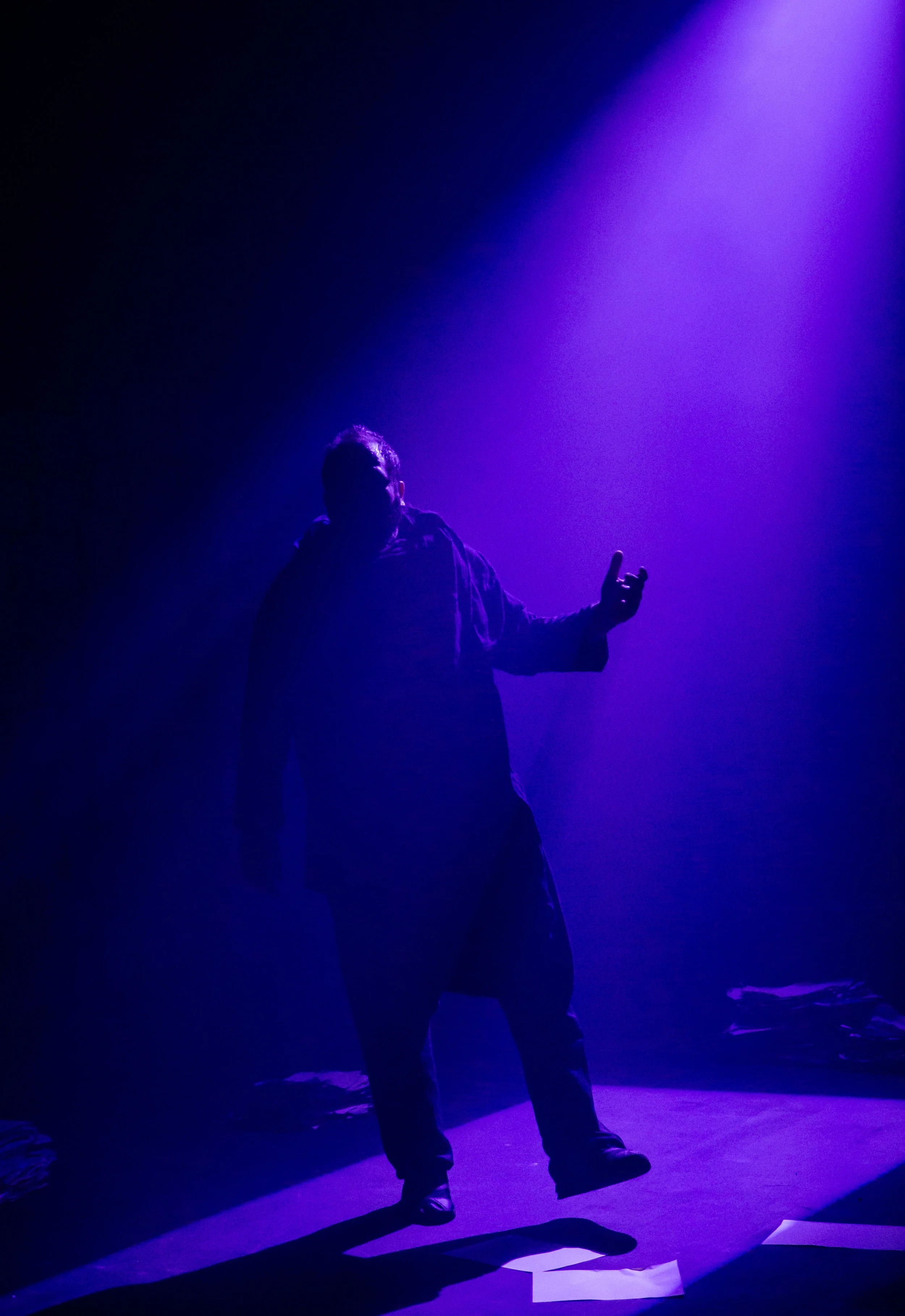 Silhouette of a person standing on a stage illuminated by purple light, with papers scattered on the floor.