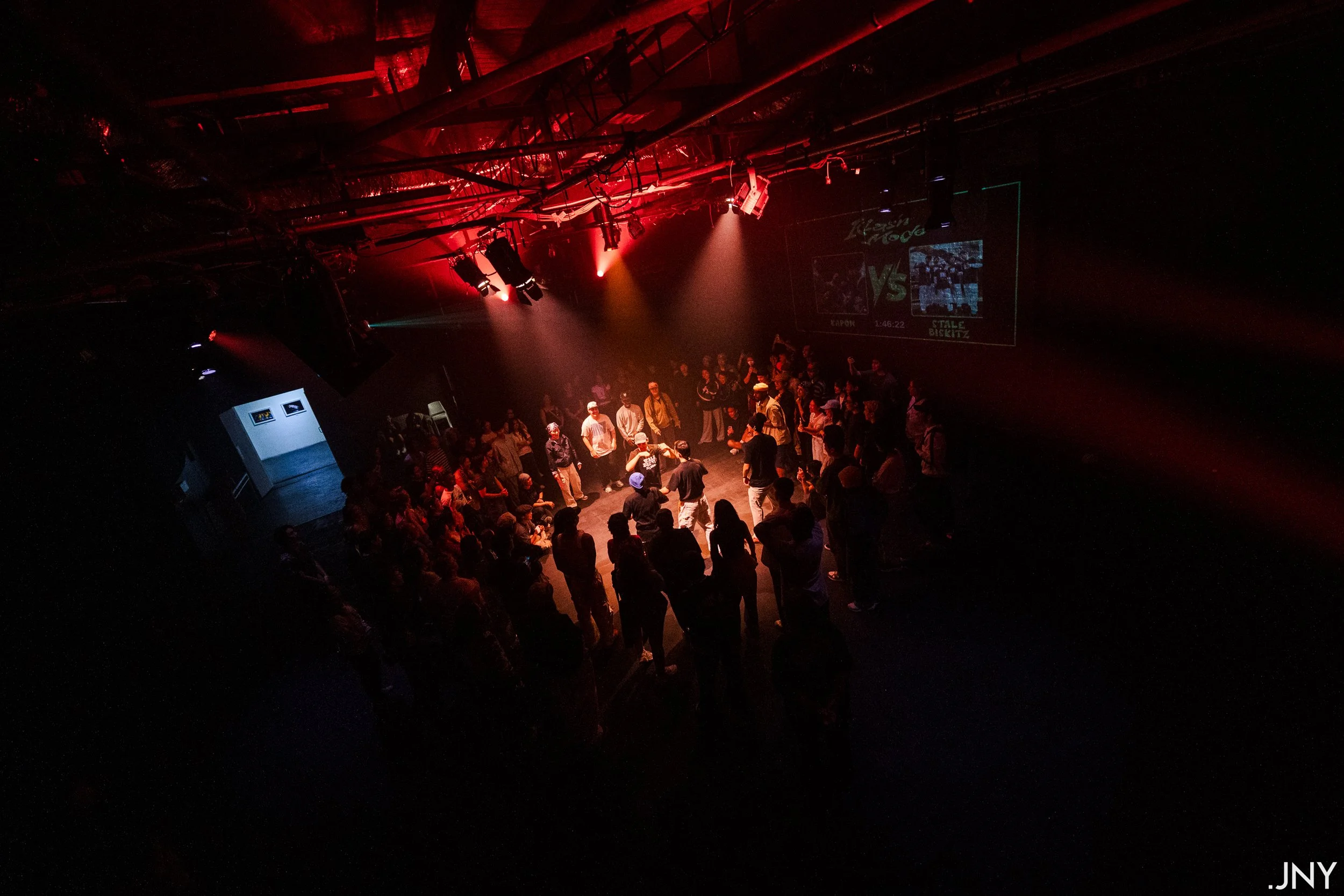 A crowd of people dancing and socializing in a dark, red-lit nightclub or concert venue. The scene includes a dance floor with some individuals wearing hats, and a large screen displaying images and text in the background.