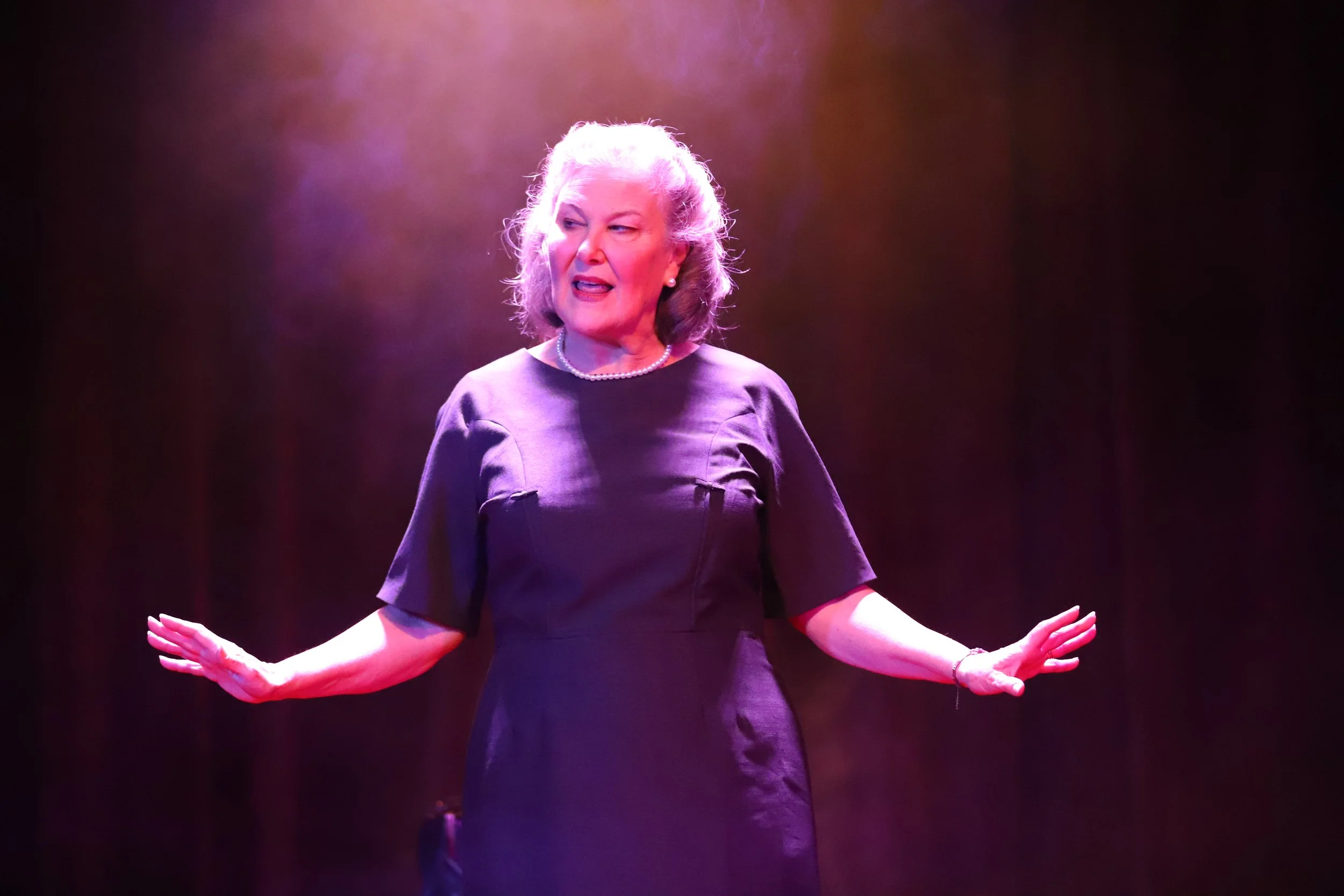 An elderly woman with gray hair, wearing a black dress, pearl necklace, and bracelet, is standing on stage with her arms extended, speaking or performing under purple stage lighting.