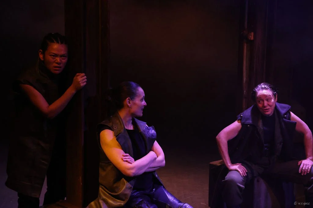 Three women in dark clothing on a stage, engaging in a theatrical performance. One woman is standing and peering from behind a wall, the second is sitting with arms crossed, and the third is seated on a box, speaking to the others.