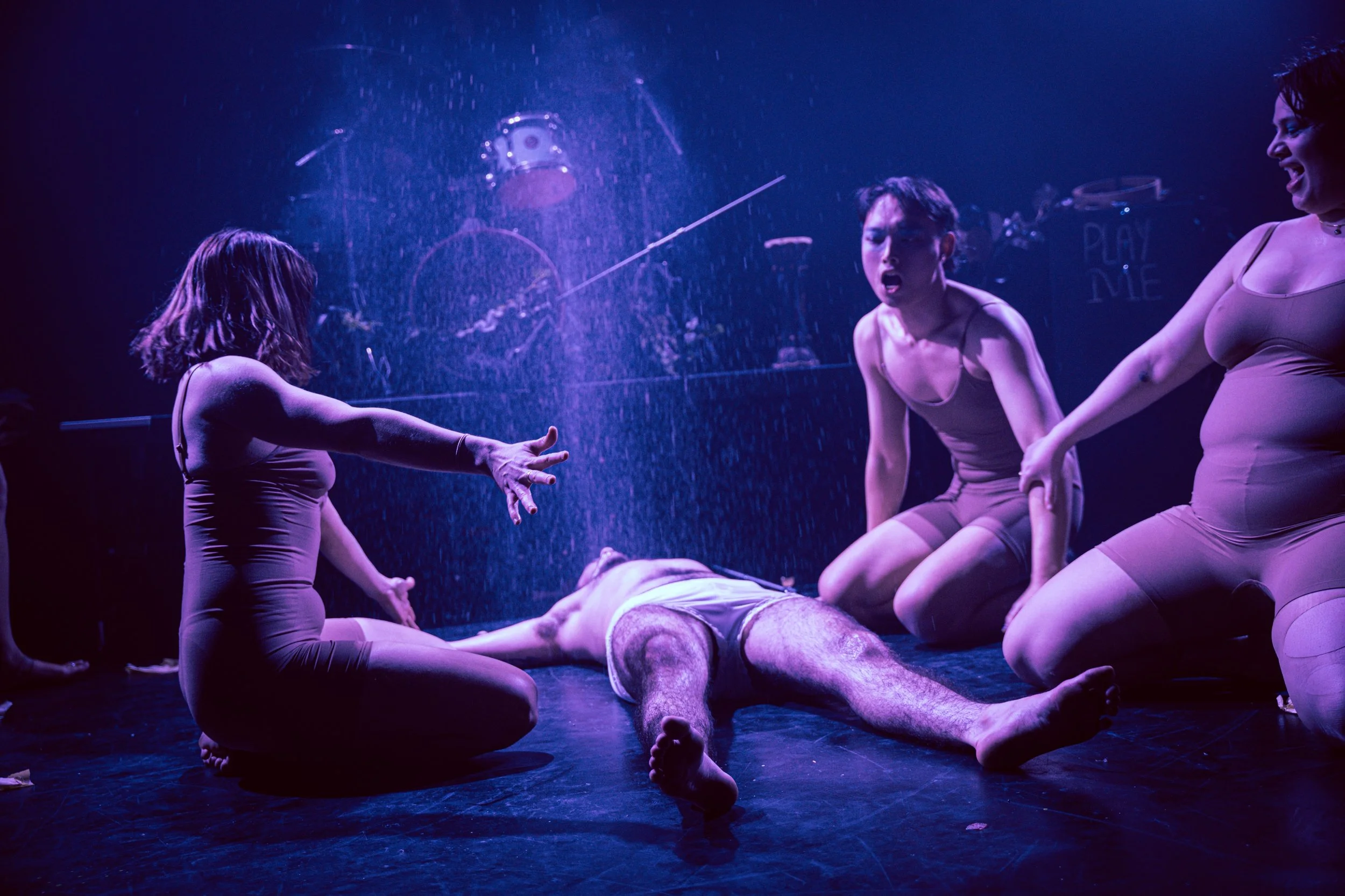 The image shows a theatrical scene with four women dressed in beige costumes surrounding a man lying on the floor. The scene is illuminated with purple lighting and water, giving a dramatic atmosphere.