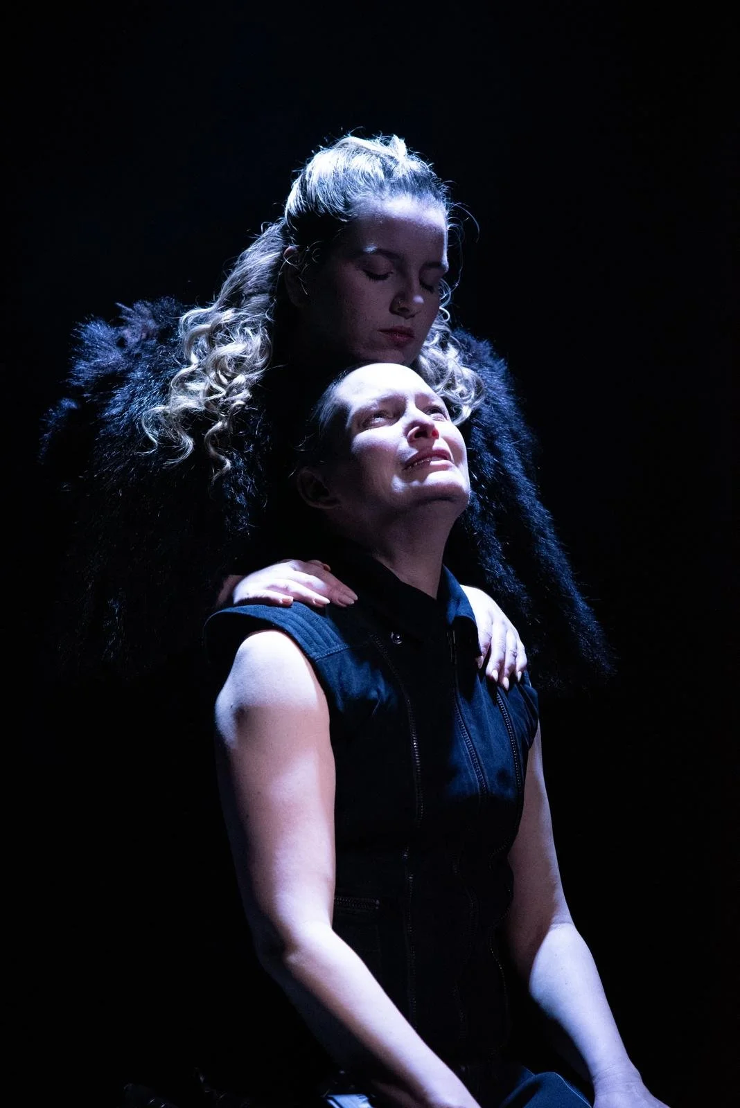 Two women are in a dark setting with dramatic lighting. The woman in the foreground has short hair, is wearing a sleeveless top, and appears emotional. The woman behind her has long, curly blonde hair, is dressed in a dark fur-lined coat, and is gent
