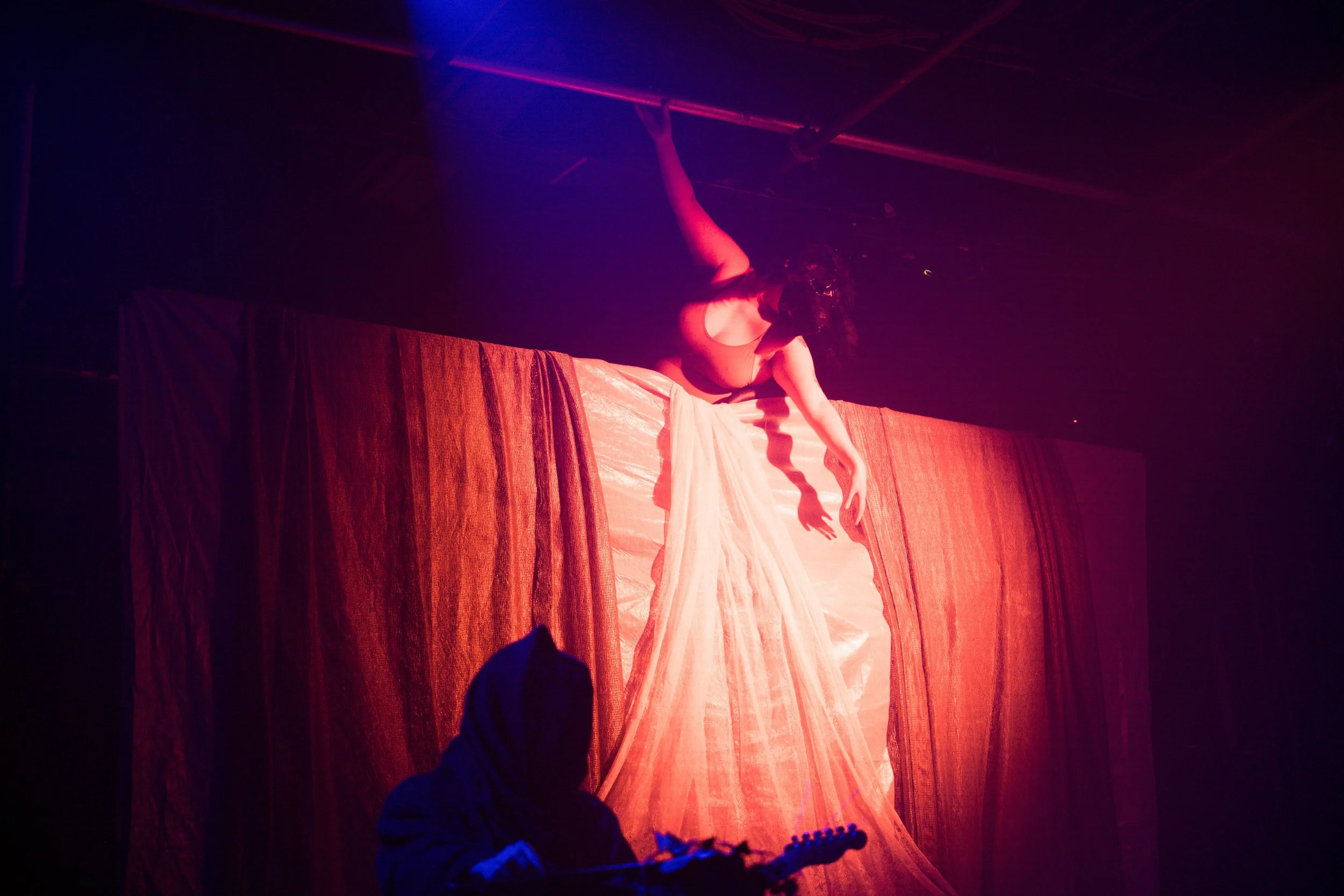Performer in a mask and flowing dress hanging upside down on a pole during a stage performance with colorful lighting.