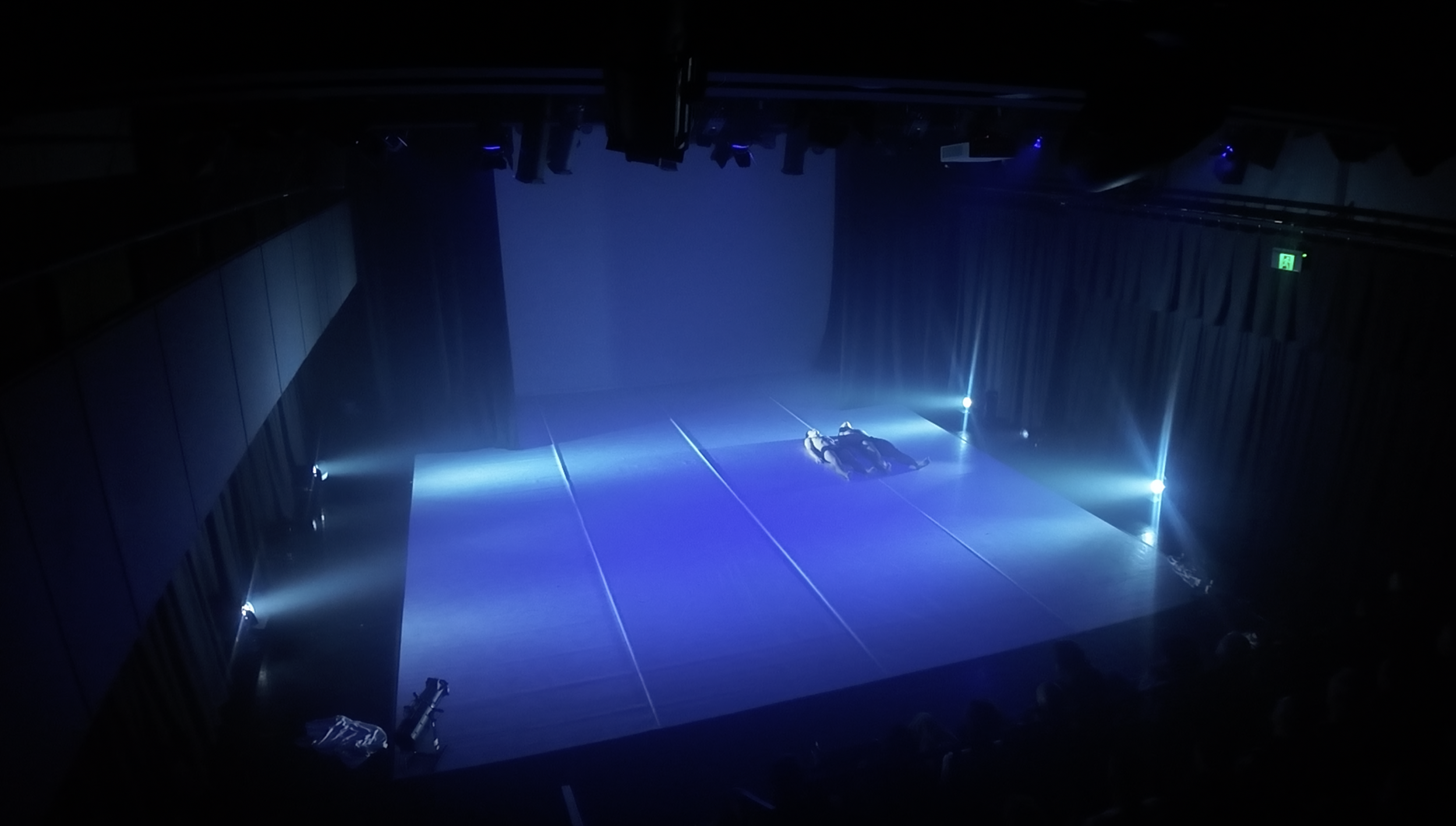 A stage with a single spotlight illuminating a person lying on the ground, with various equipment around, in a dark theater or auditorium.