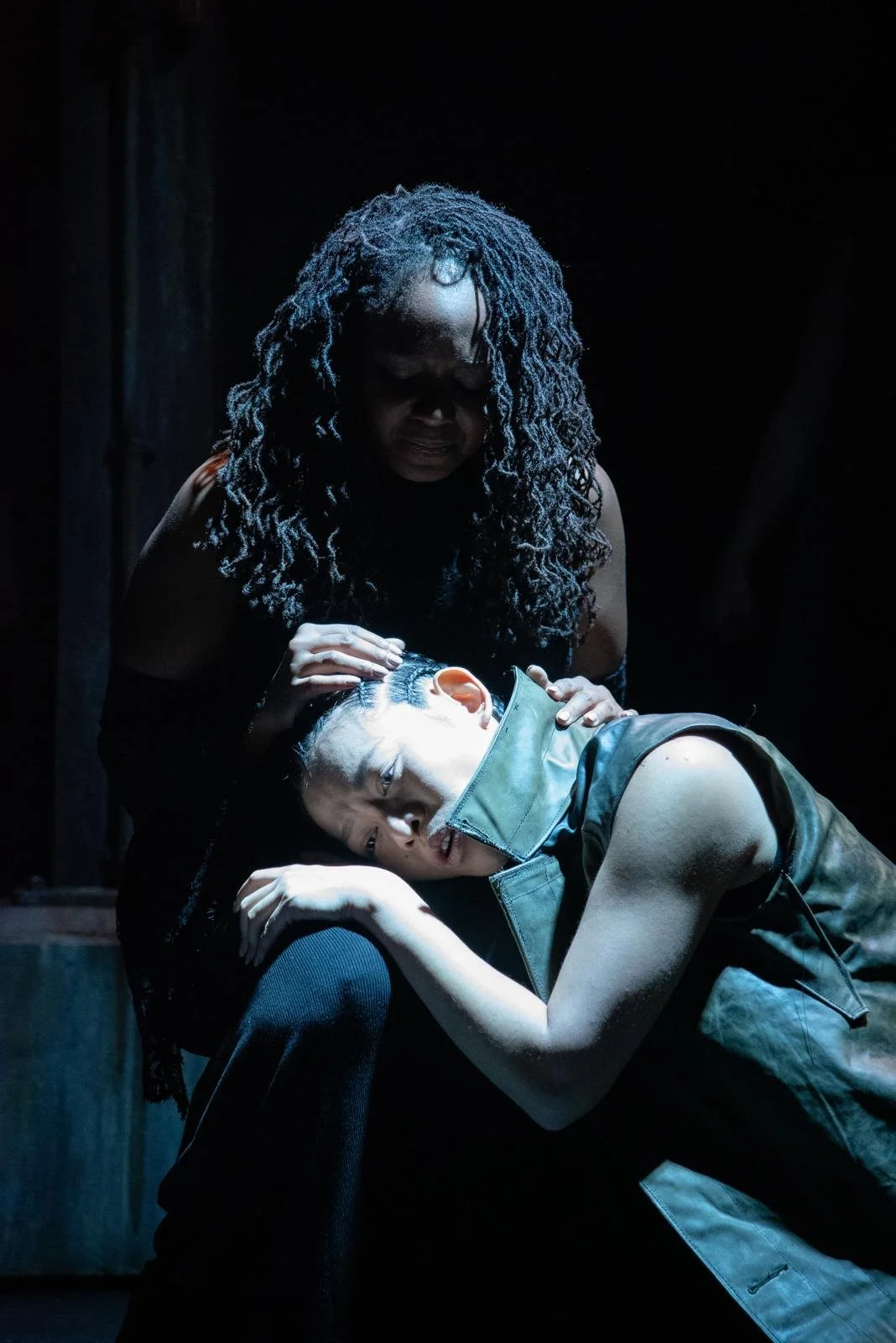 A woman with curly hair, wearing glasses, is leaning over a person resting their head on her lap. The resting person has styled hair, a sleeveless leather vest, and is looking up with a serious expression. The scene is dark with focused lighting high