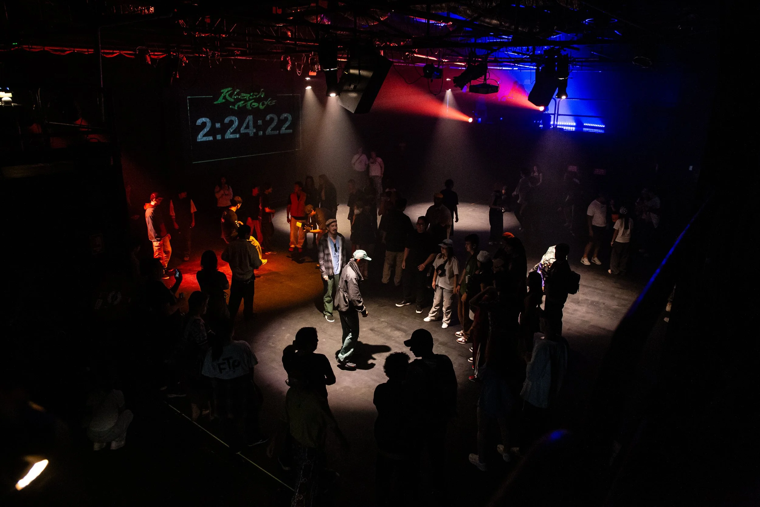 People dancing and socializing in a dark, lit stage or club with colored overhead lights and a digital clock display reading 2:24:22