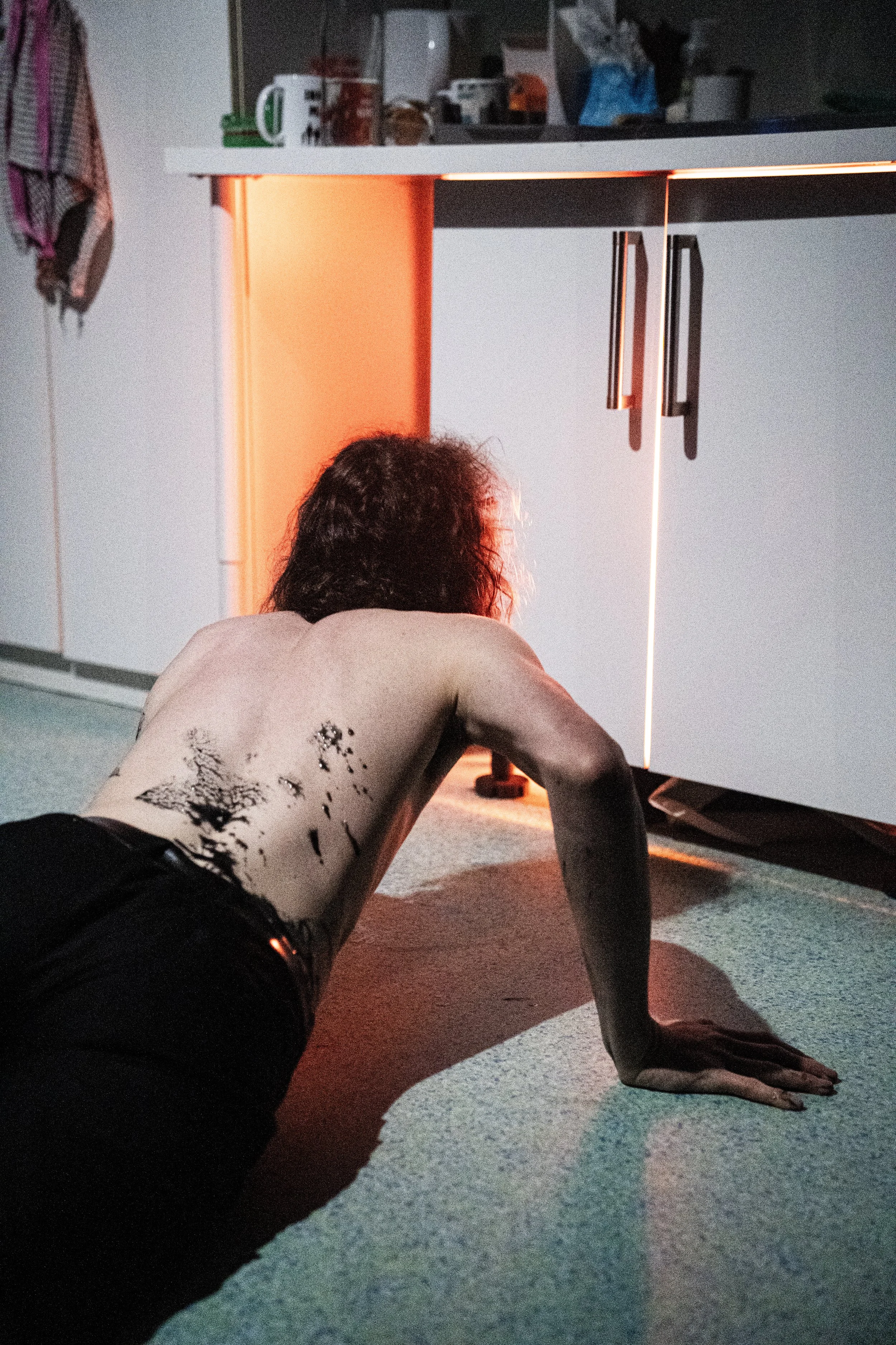 Person with curly hair, shirtless, crawling on the carpeted floor in a kitchen with a small orange light underneath the cabinet.