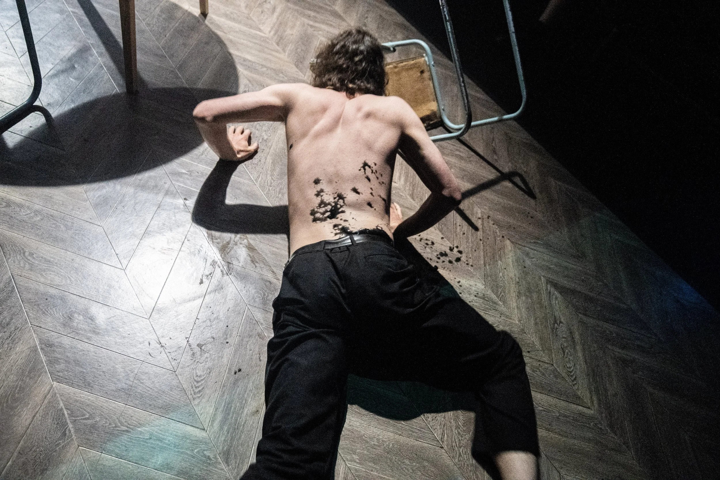 A shirtless young man with brown hair lying on a wooden floor with black paint splattered on his back, next to a small stool and a black surface, with shadows cast in a dimly lit room.