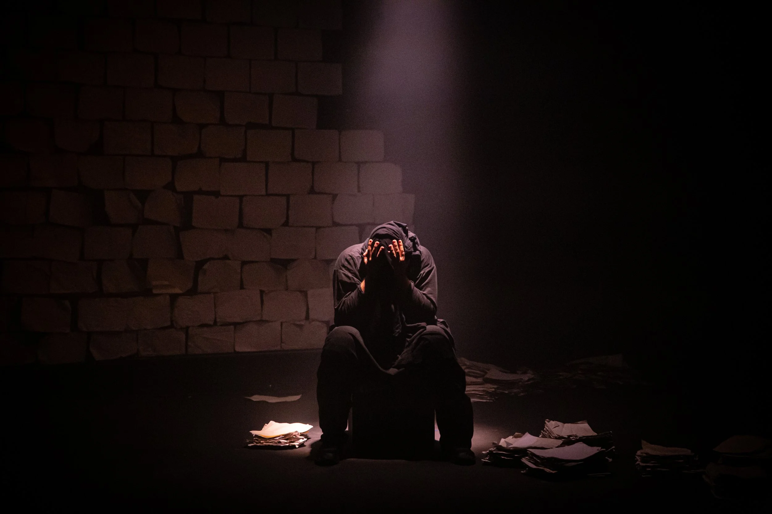 A person dressed in black sitting on a box, with hands covering their face, in a dimly lit space with a brick wall background and piles of papers on the ground.