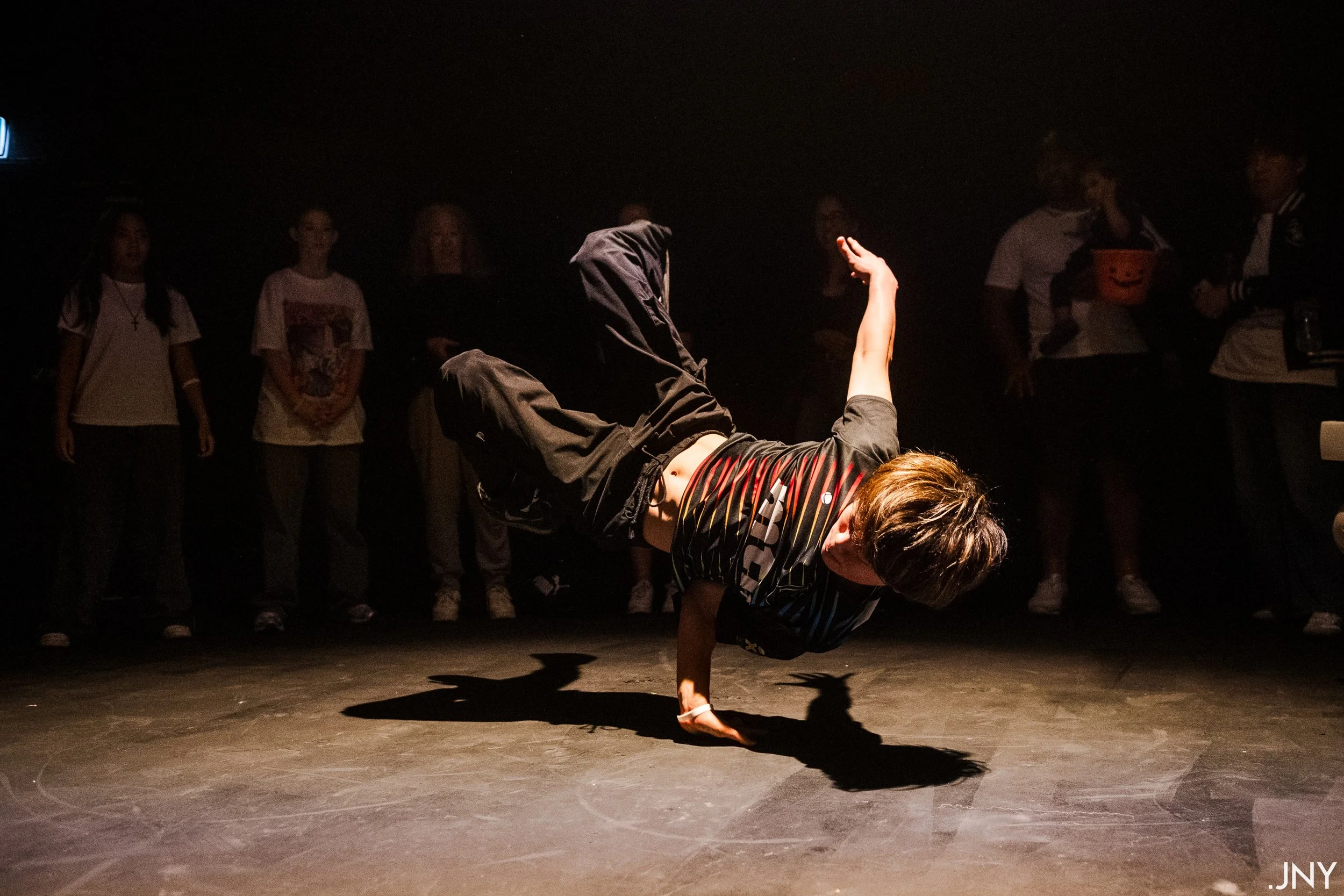 Person performing a breakdance move on a dark stage with a spotlight, while a group of people watch in the background.