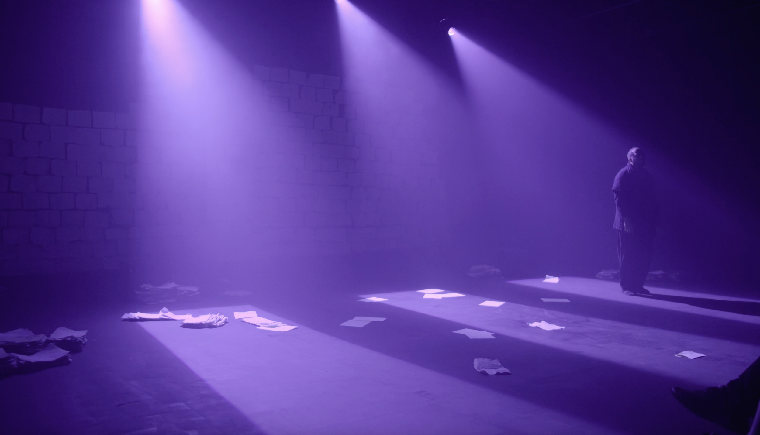 Dimly lit stage with purple lighting, scattered papers on the floor, a lone person standing in the shadows on the right, and a brick wall in the background.