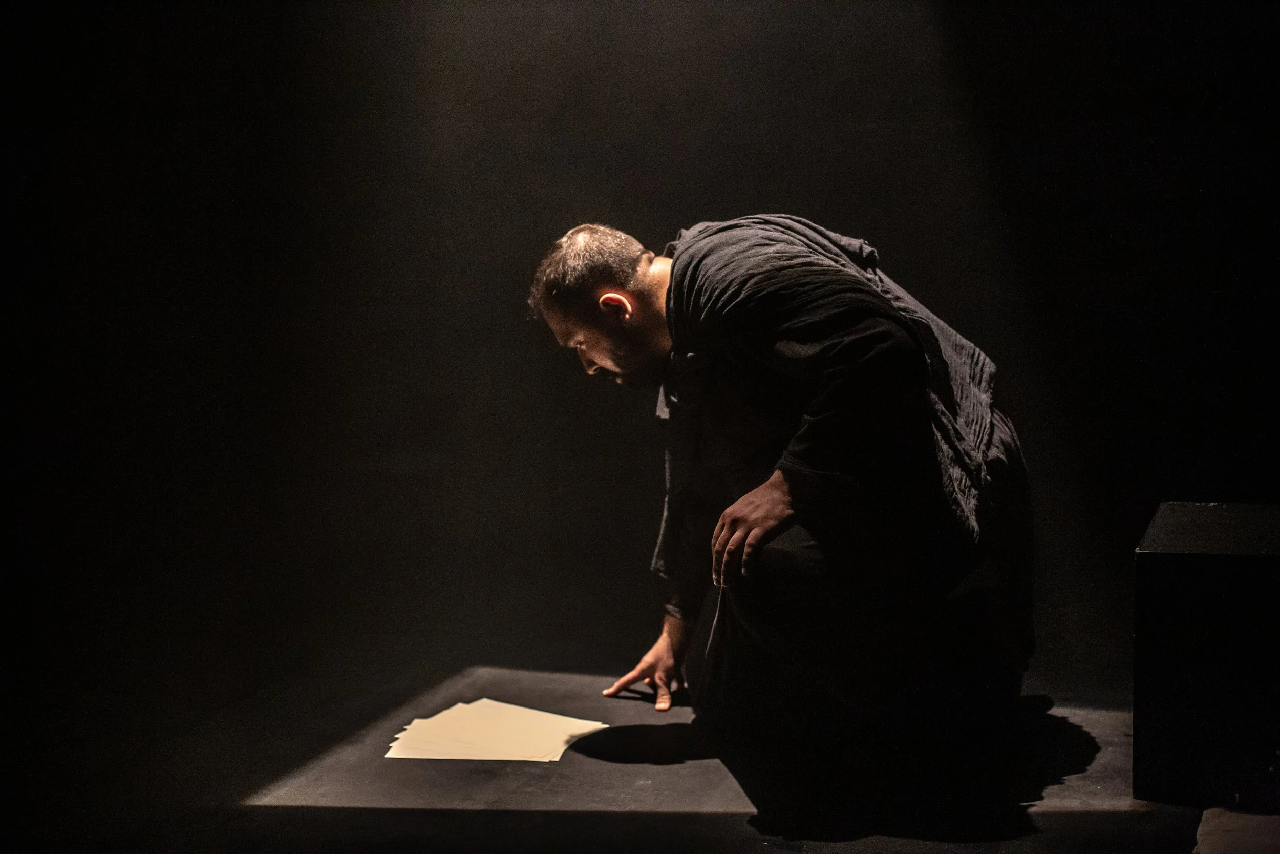 A man kneeling on a dark stage in low light, looking down at a stack of papers on a black surface in front of him.