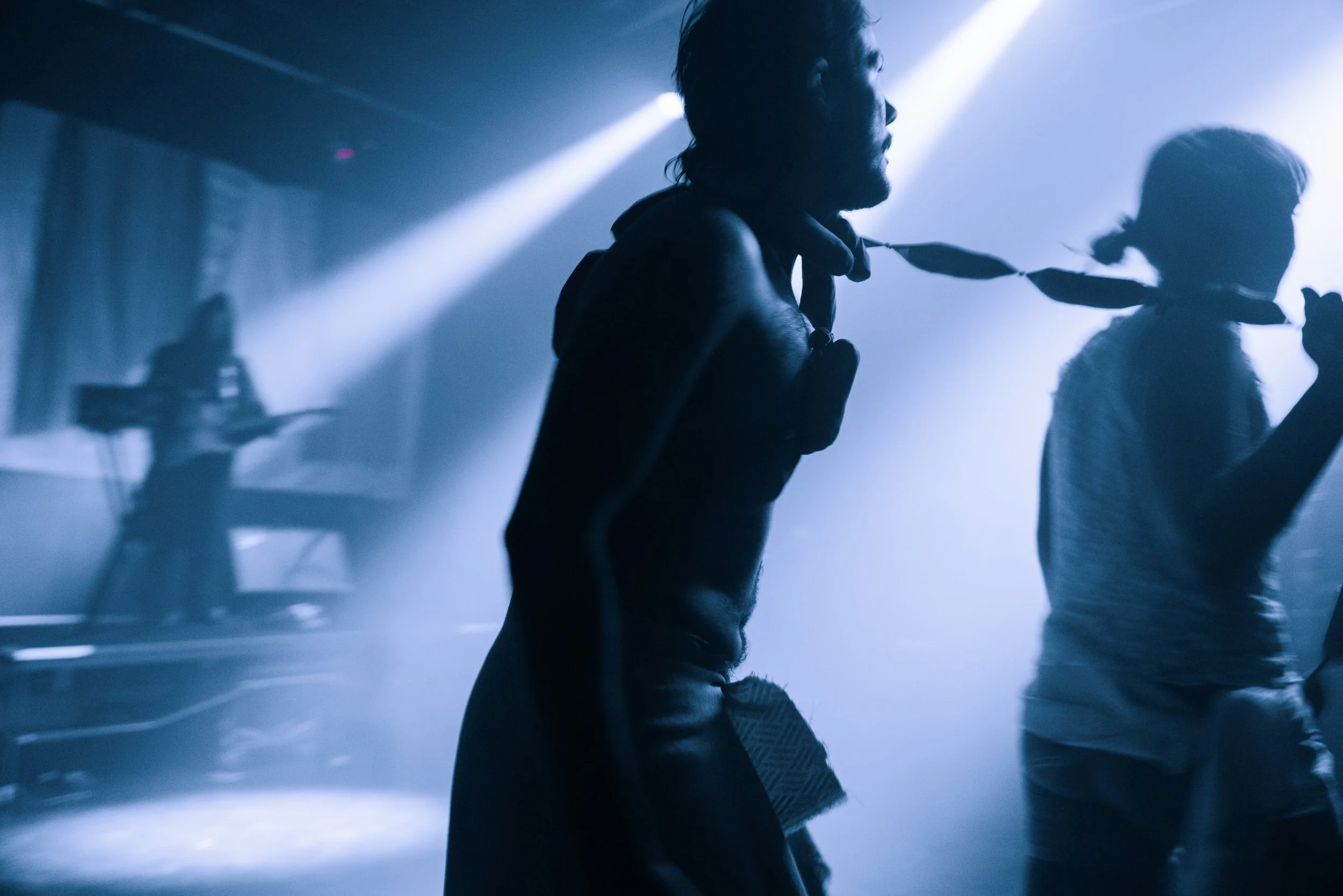 Silhouettes of two people on stage, with one person wearing a scarf, and a DJ in the background with a turntable, under blue stage lighting.