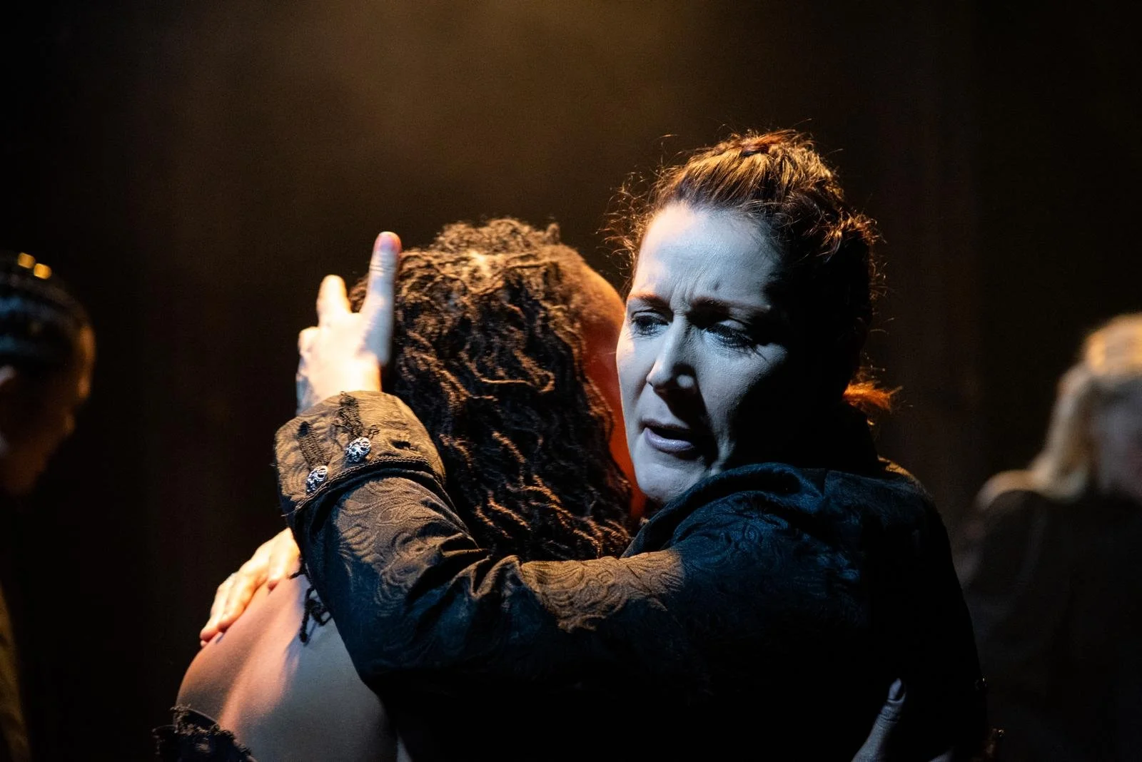 A woman with dark hair pulled back holds another person closely, comforting them. The scene is dark and dramatic with warm lighting.