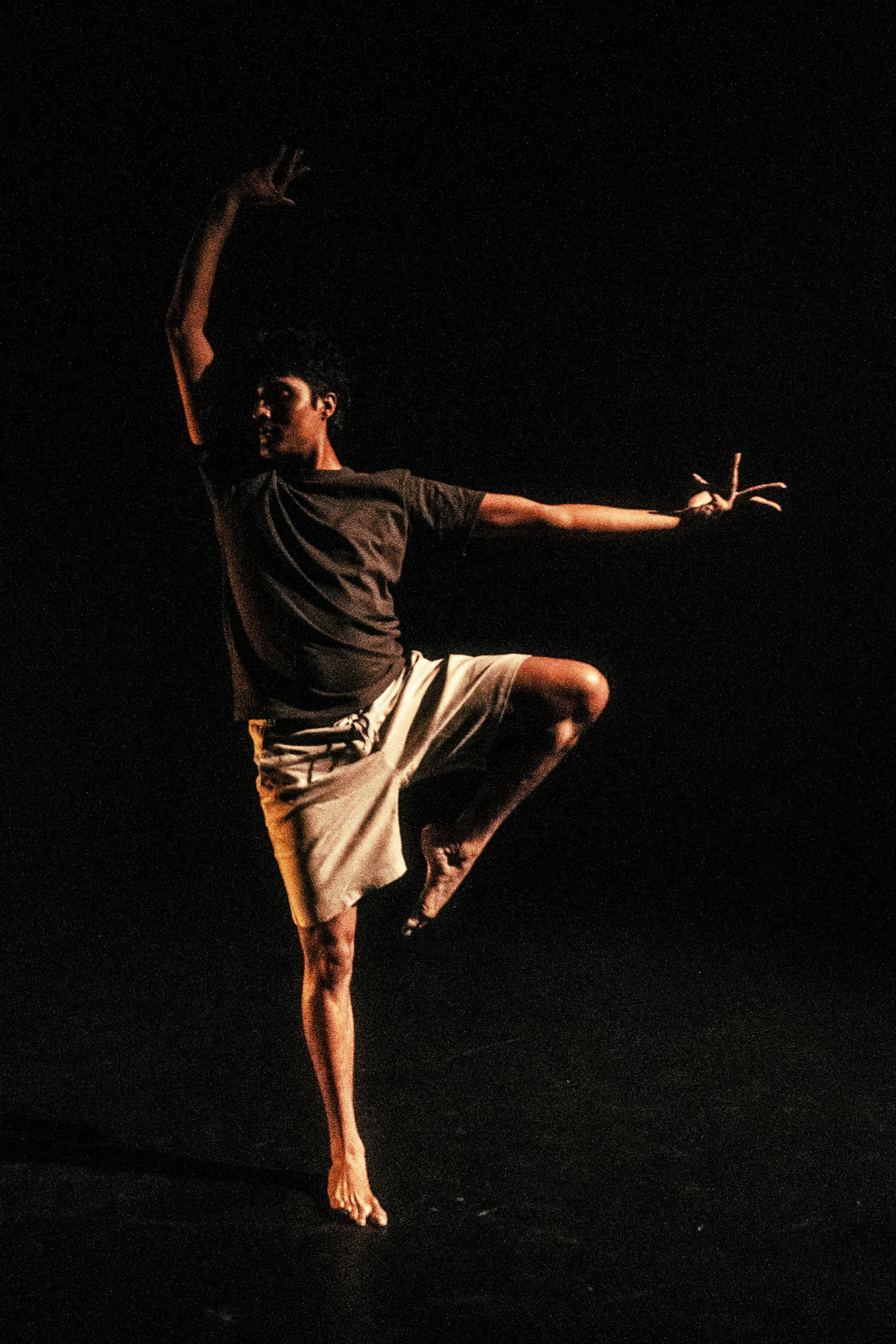 A man dancing on stage in a dark background, balancing on one foot with arms raised and in a dynamic pose.