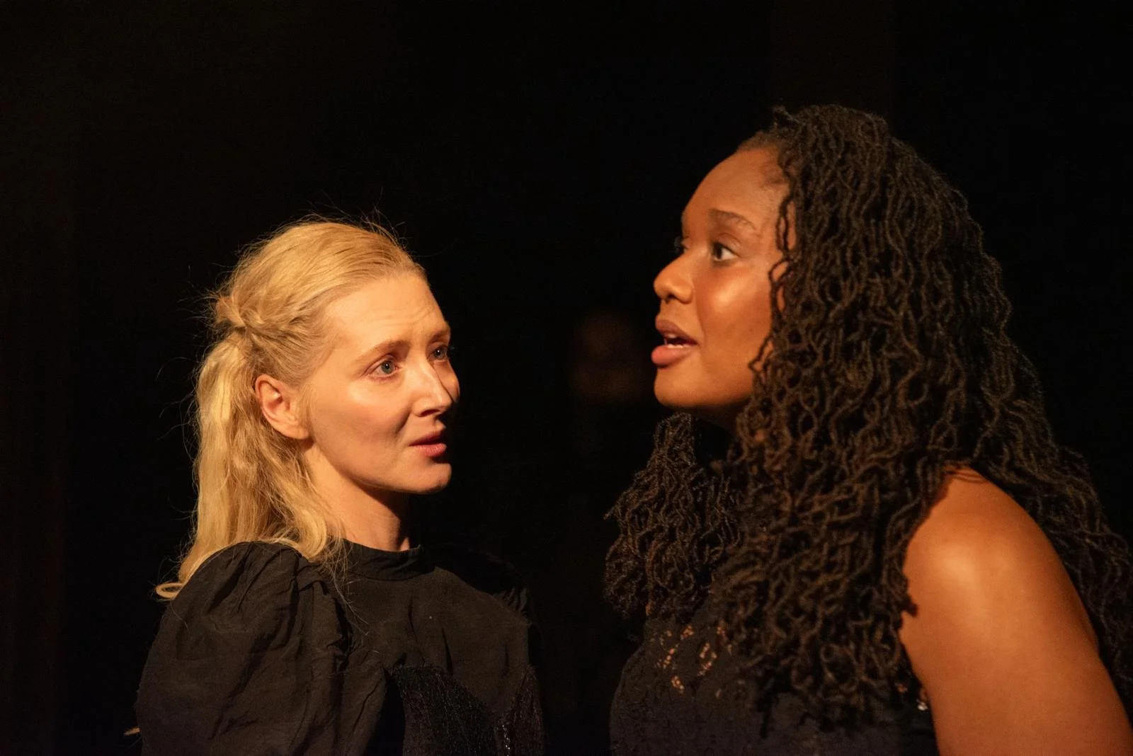 Two women face each other closely, one with blonde hair and the other with dark, curly hair, both expression serious, in a dark setting.