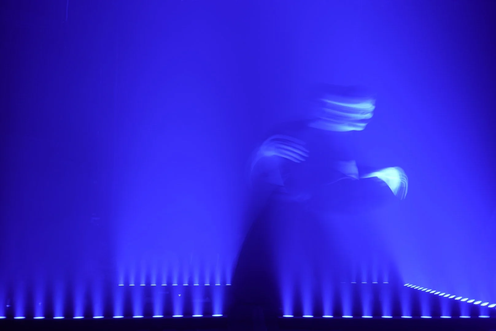 A person sitting on the ground in front of a large illuminated blue water fountain display, with their face illuminated by the blue light.