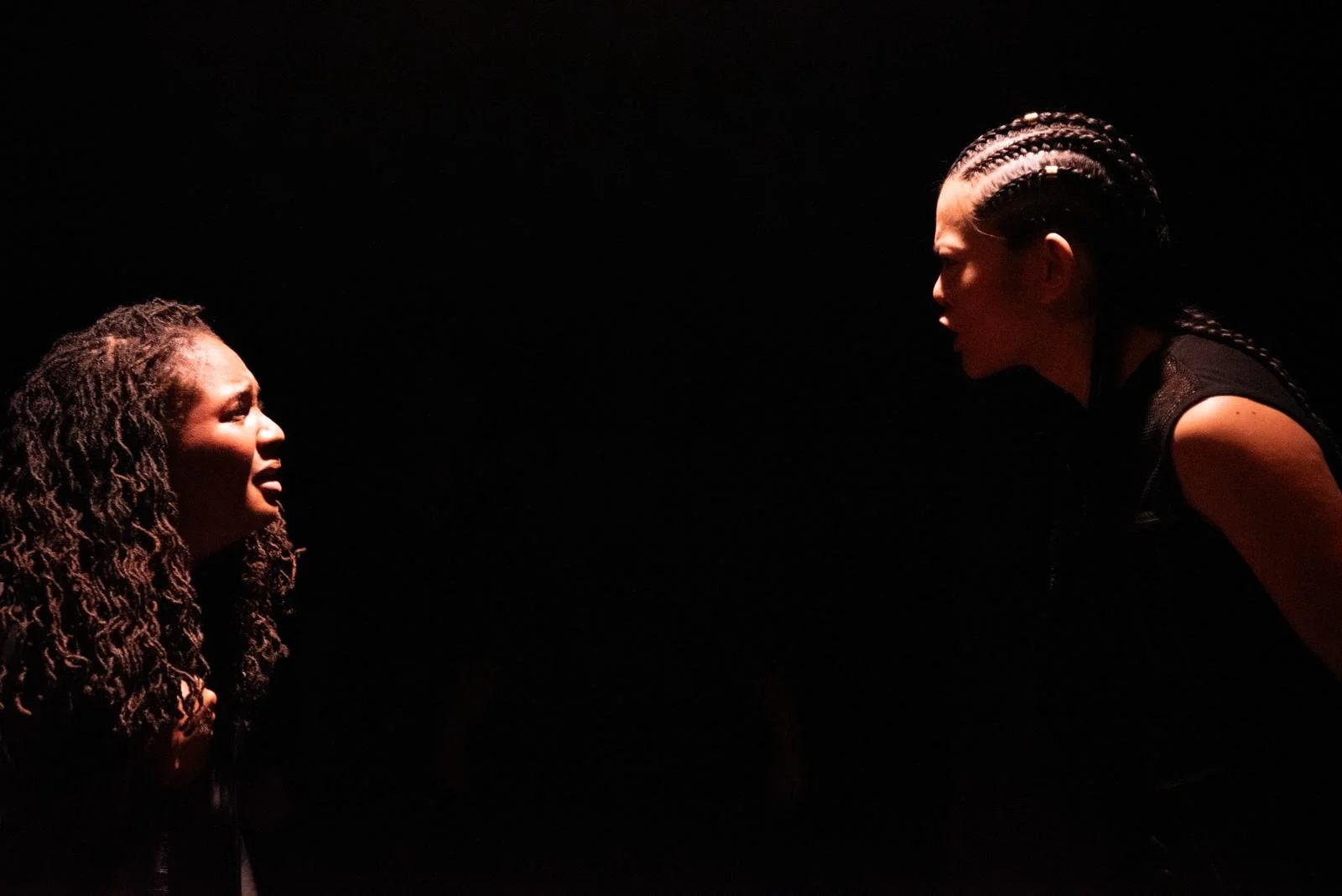 Two women facing each other in dramatic light against a dark background, one with curly hair and the other with braided hair, appearing to be in an intense conversation or confrontation.