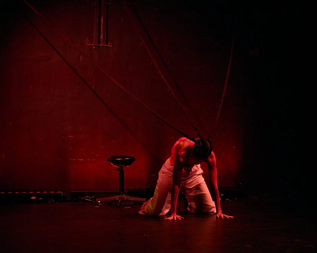 Performer with braided hair wearing light-colored pants, crouched on stage with head down, illuminated by red lighting, with chains hanging from the ceiling.