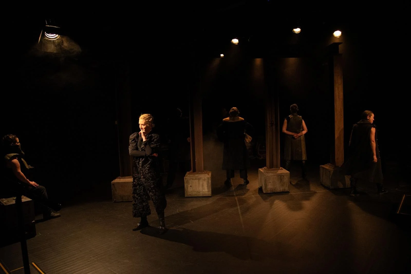 People on a dimly lit stage with wooden pillars, some standing and one sitting, in a theatrical scene.