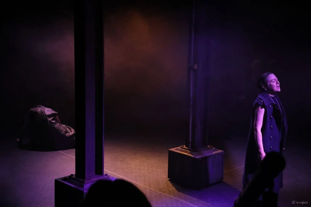 A woman singing on stage under purple lighting, with a bag on the floor in the background.