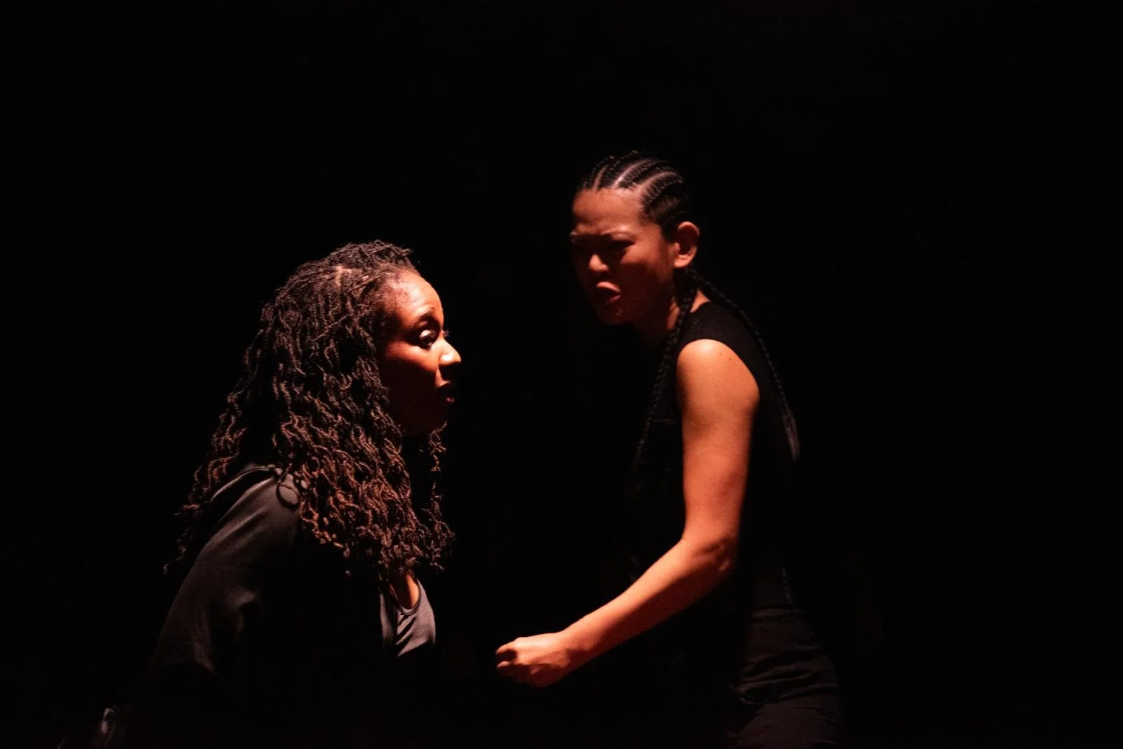 Two women engaged in an intense conversation, one with long curly hair and the other with braided hair, illuminated against a dark background.