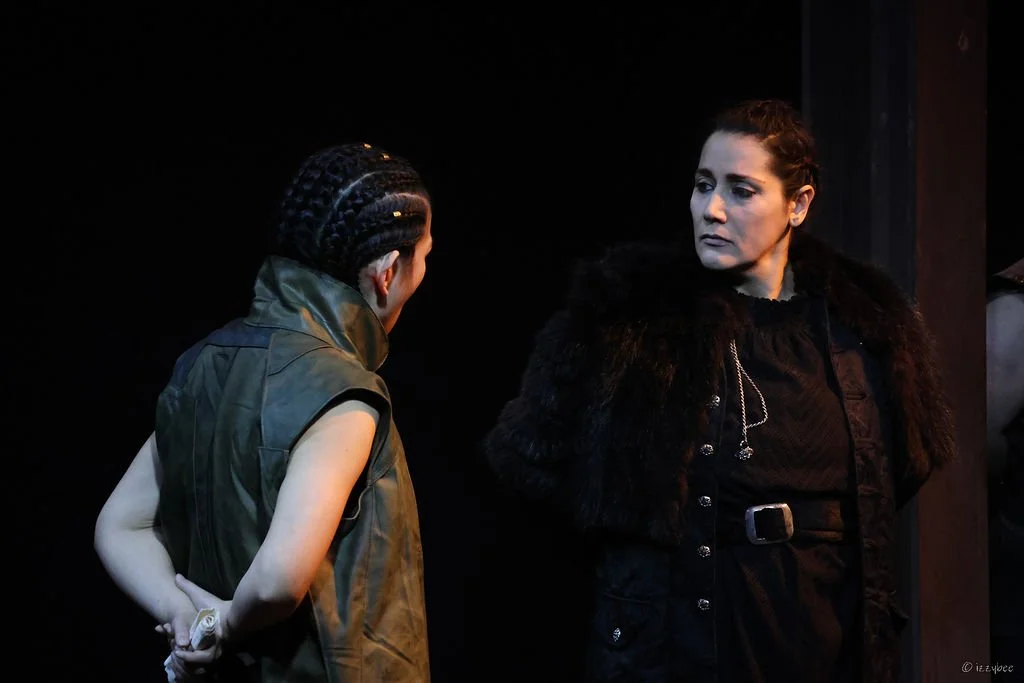 Two women having an intense conversation on stage. One is wearing a sleeveless green vest with braids, the other is dressed in a black coat with fur trim.