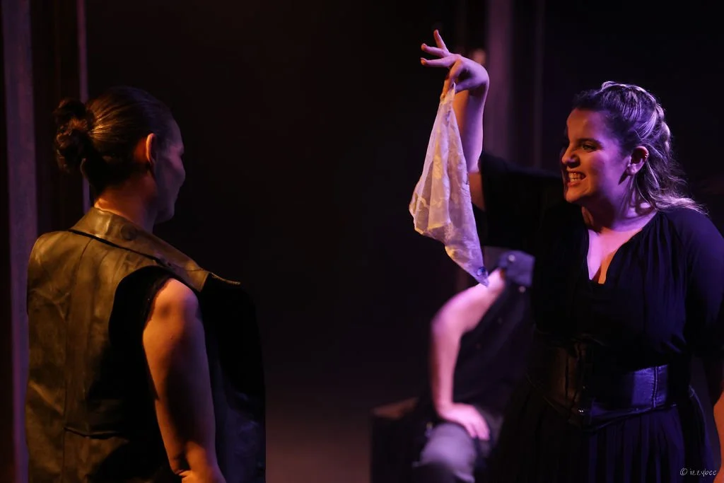 Two women engaging in a theatrical performance on stage; one woman is smiling while the other looks at her, with dark background and purple lighting.