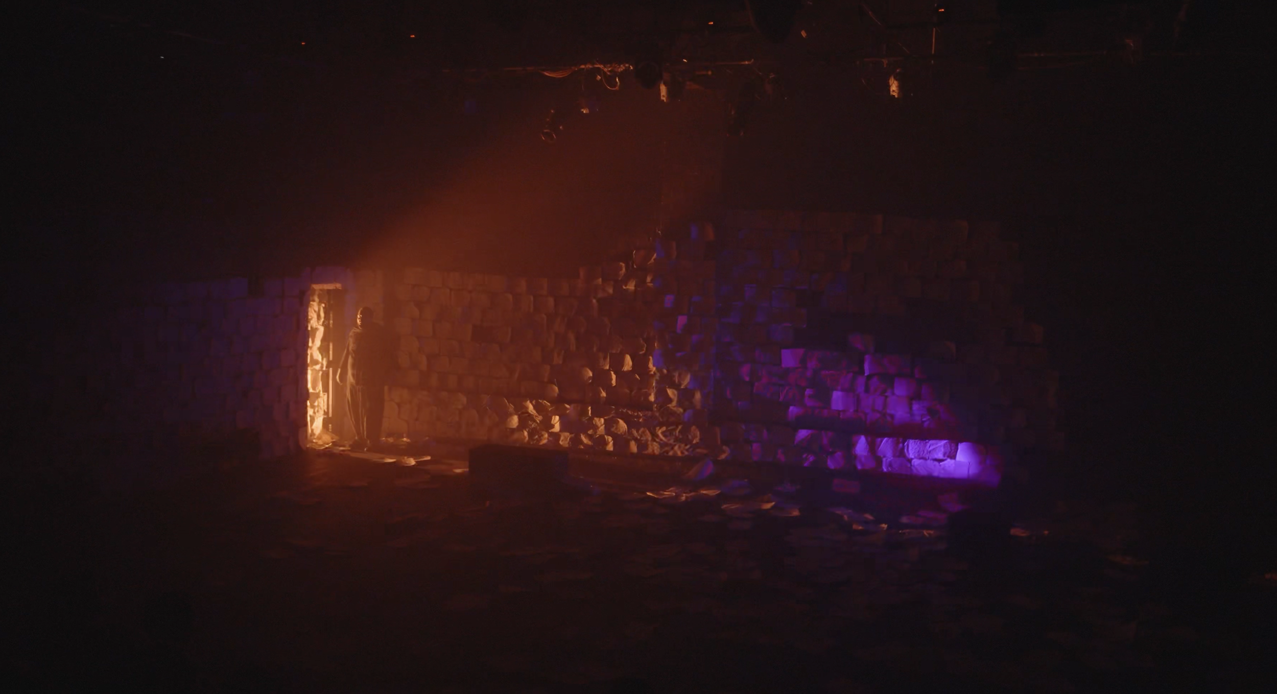 A person standing in a doorway lit with orange and purple lights, in an indoor space with a stone wall and scattered papers on the floor.