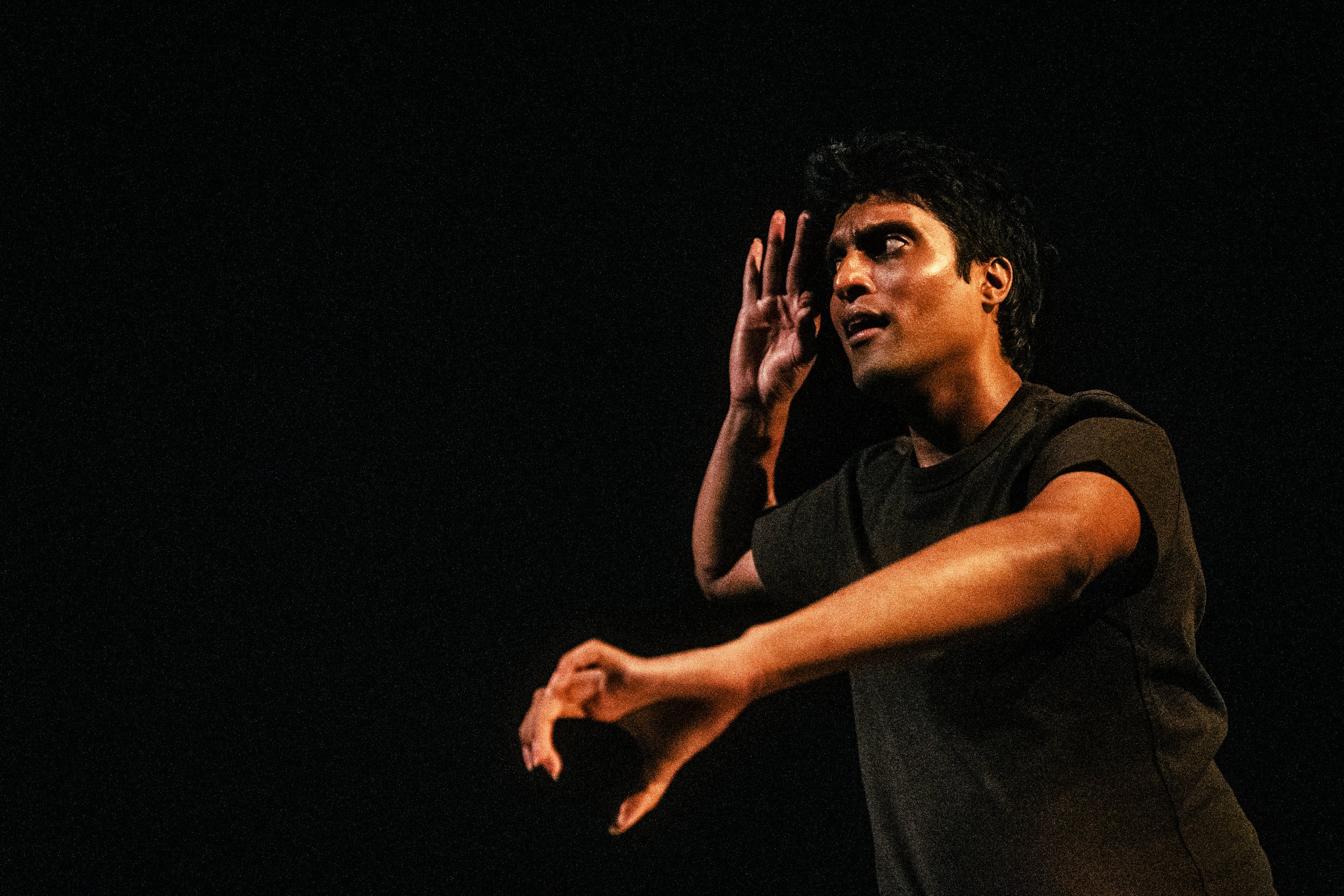 A young man with dark, curly hair wearing a dark T-shirt, performing a theatrical gesture against a black background, with his hand near his ear and his mouth slightly open.