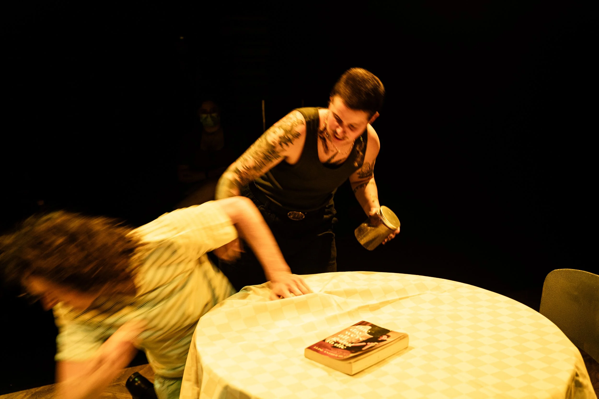 A woman with tattoos on her arms is standing over a person in yellow striped pajamas, grabbing their arm during a confrontation, with a book on the table and a dark background.