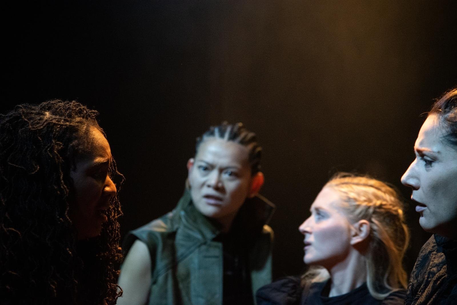 Four women with intense expressions in a dimly lit setting, facing each other in a serious conversation.