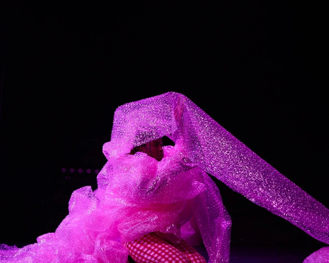 Person wearing an iridescent pink outfit with a matching umbrella, illuminated against a dark background.