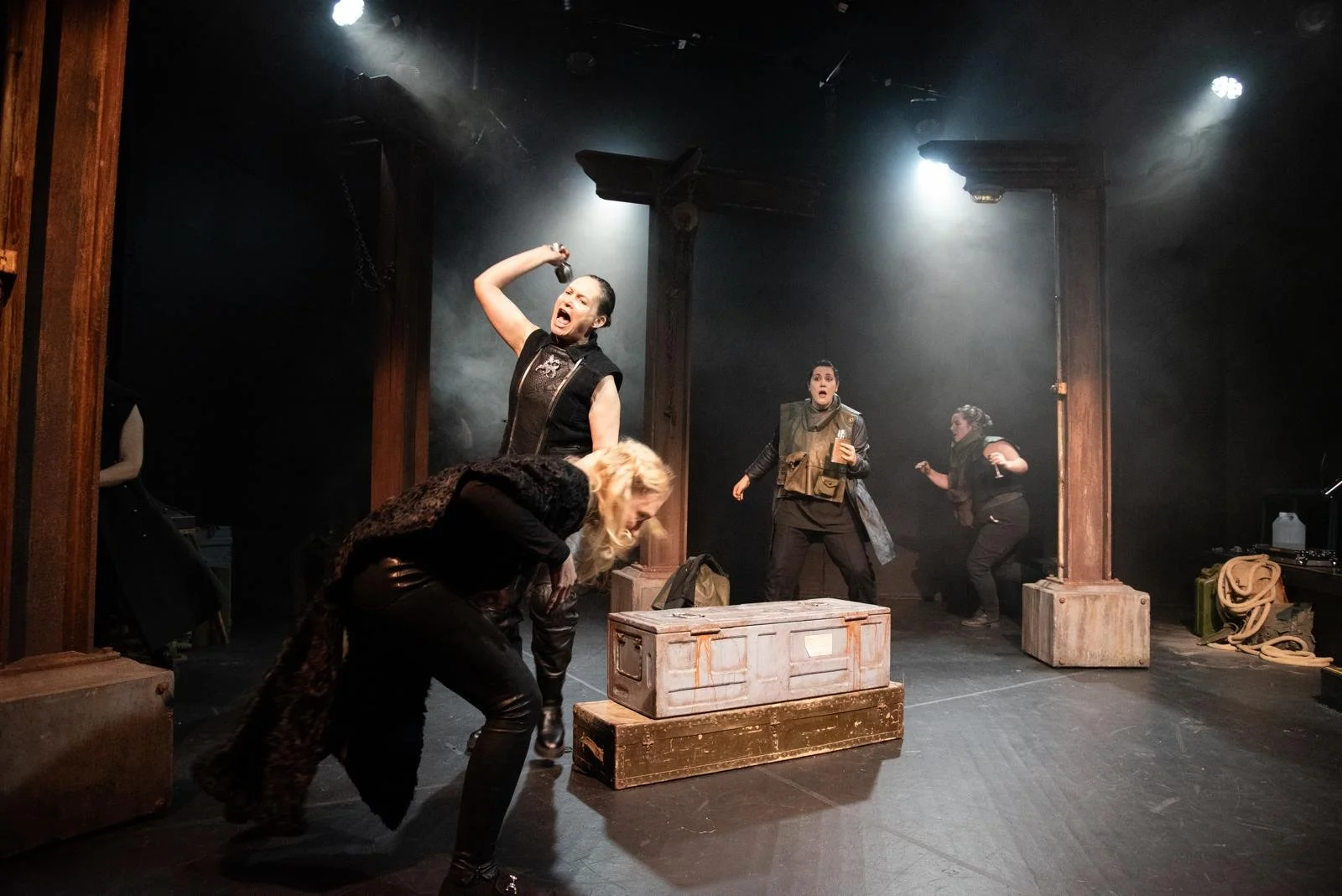 Women acting aggressively on stage, with one woman shouting and raising a hand, and others in various intense poses, amidst wooden box props and dramatic lighting.