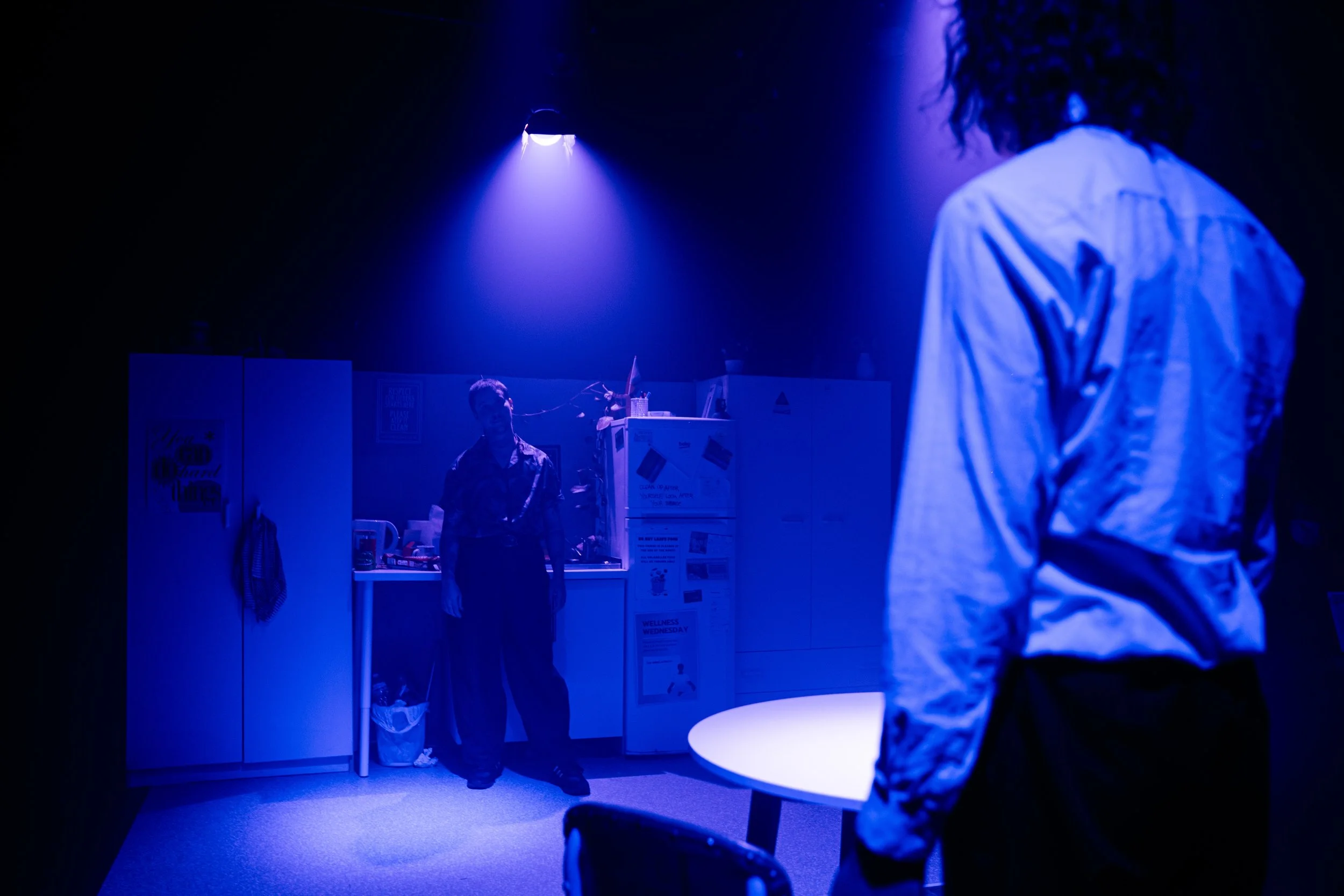 A dark room illuminated by blue light with two people, one standing near a table and the other facing away, in an office or storage room with cabinets and posters.