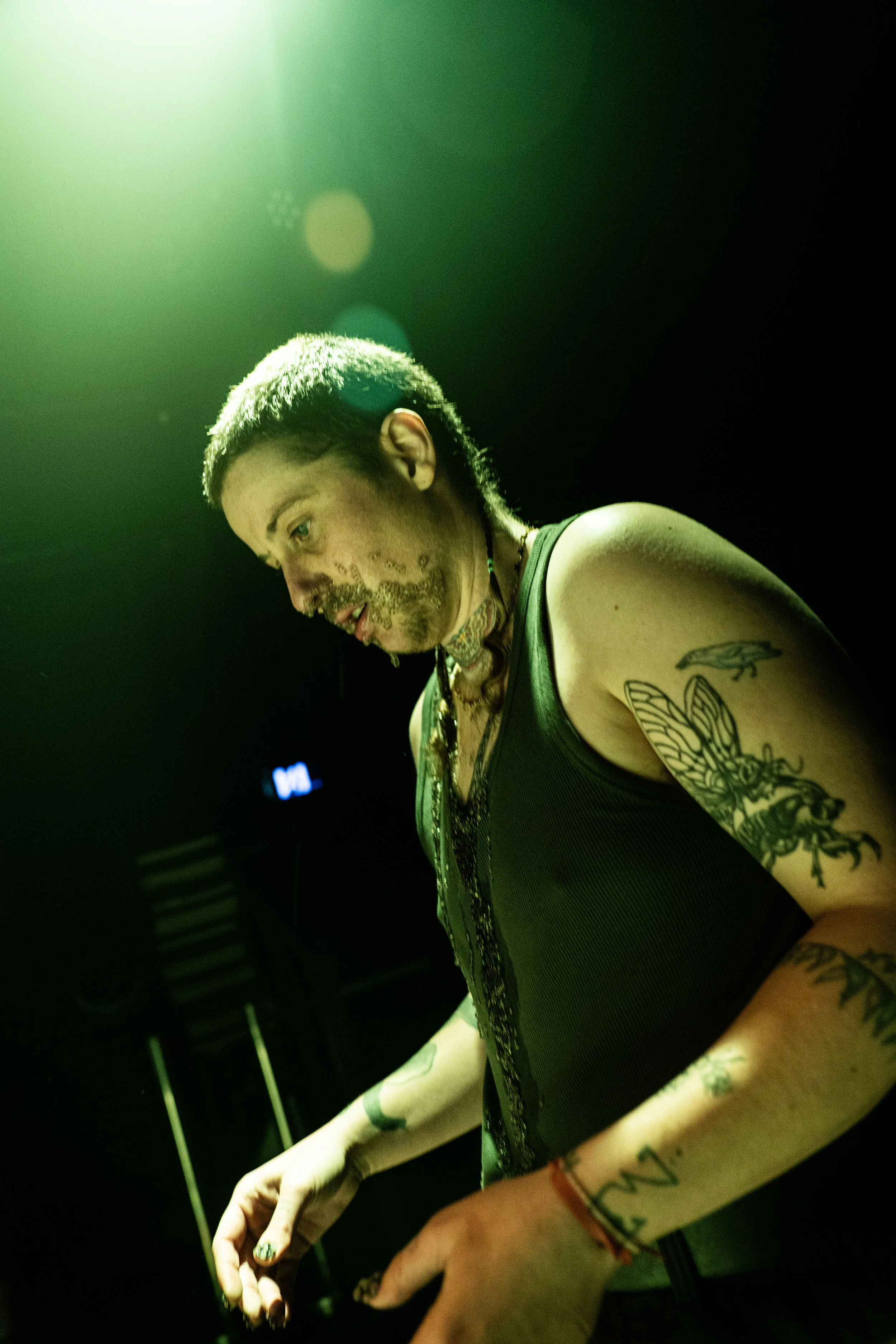 A man with short hair, tattoos on his arms, and a beard, wearing a sleeveless shirt, is looking down and smiling in a green-lit environment, possibly DJing.