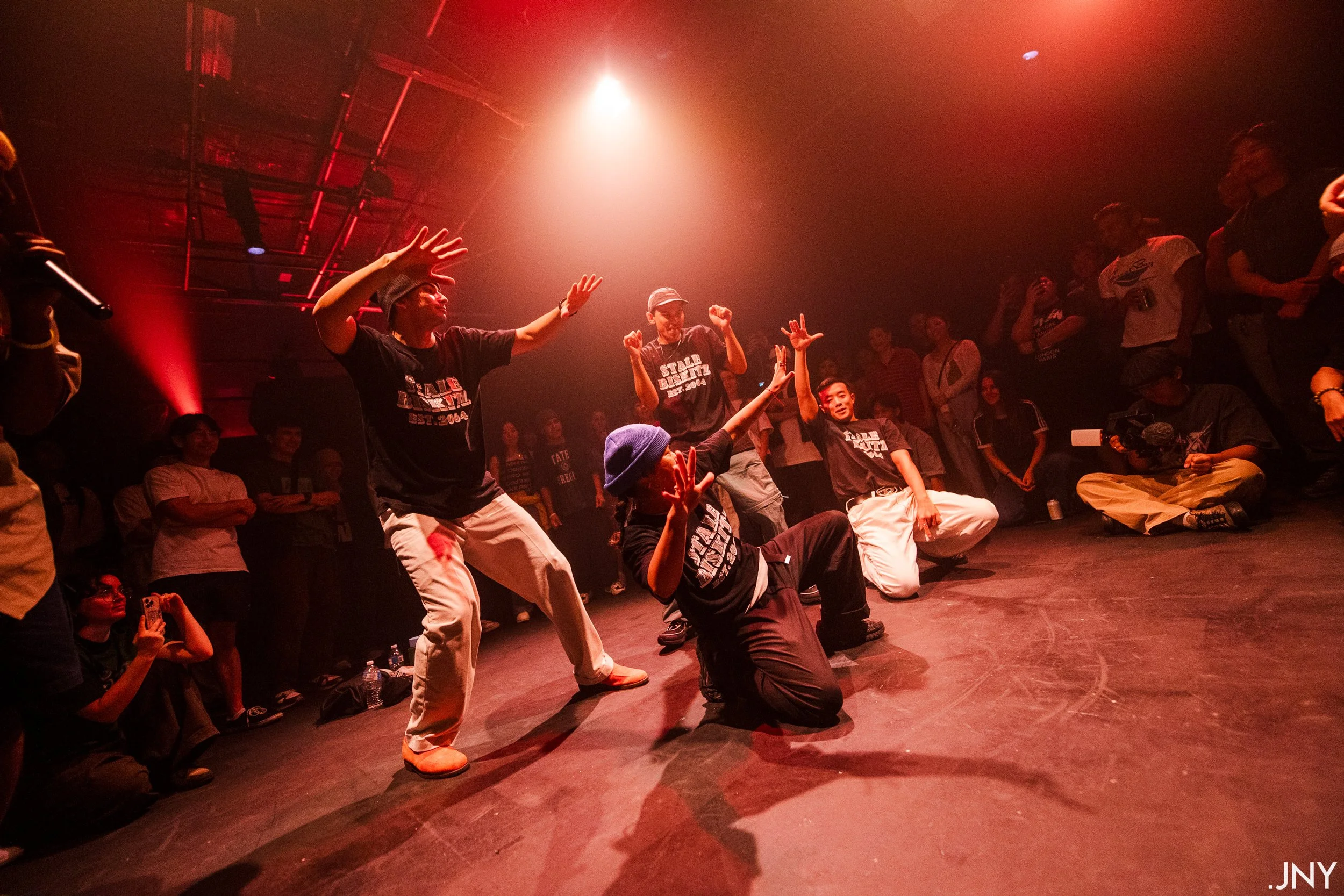 A group of performers dancing on a stage illuminated by red lighting, surrounded by an audience during a dance performance.