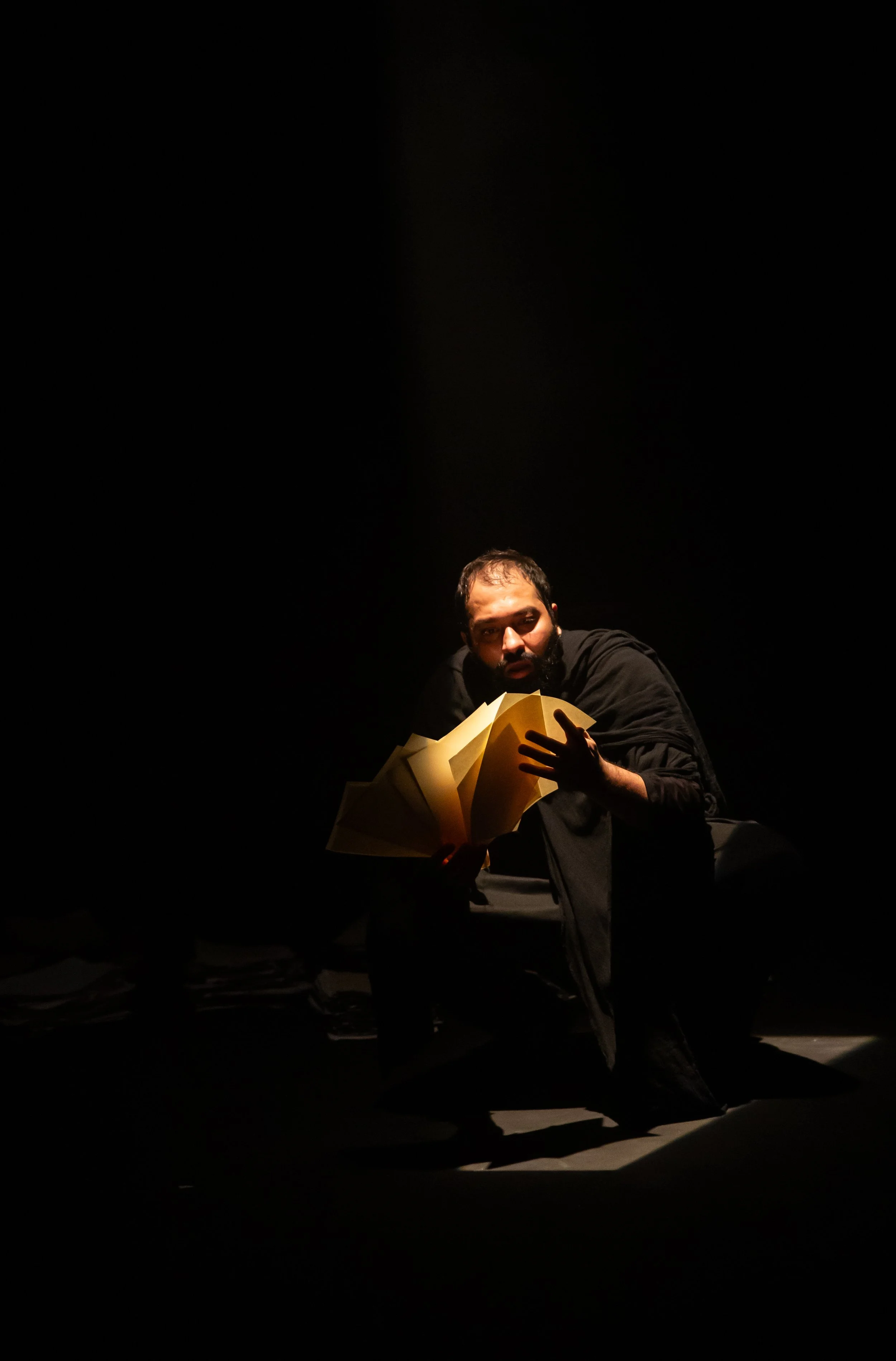 A man with a beard in a dark room, illuminated by a spotlight, sitting on the floor, holding and looking through a large open yellow book.