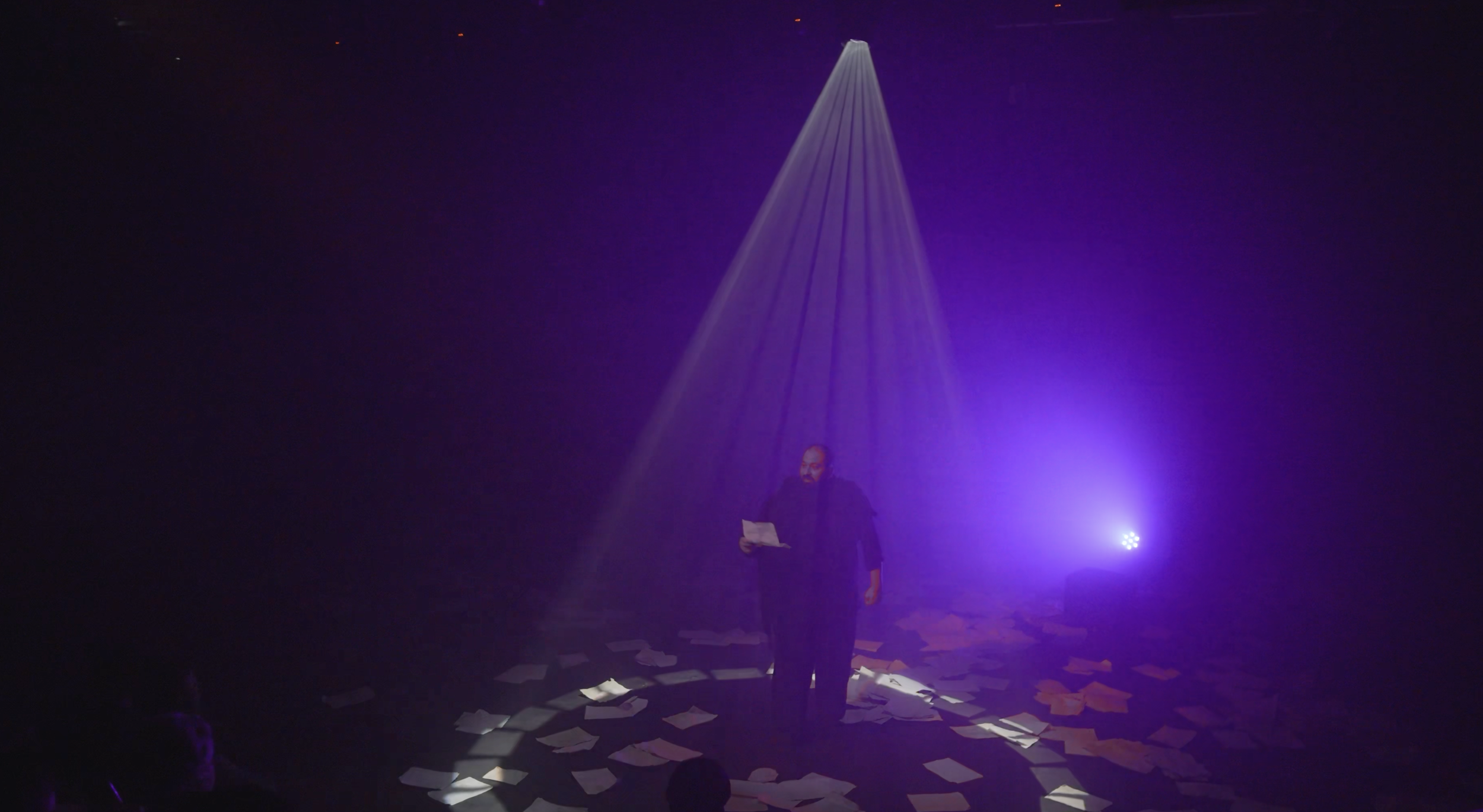A person standing under a purple spotlight on a dark stage, holding papers, with scattered papers on the floor.