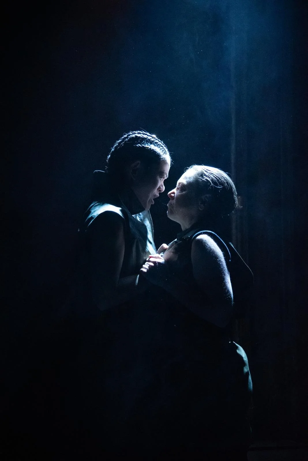 Two women face each other closely with intense expressions, illuminated by spotlight, dark background, dramatic scene.