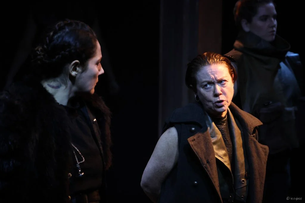 Three women engaged in a serious conversation in a dimly lit setting. The woman in the center has short hair, furrowed brows, and wears a sleeveless jacket. The woman on the left has longer hair pulled back, and the woman on the right, slightly blurr