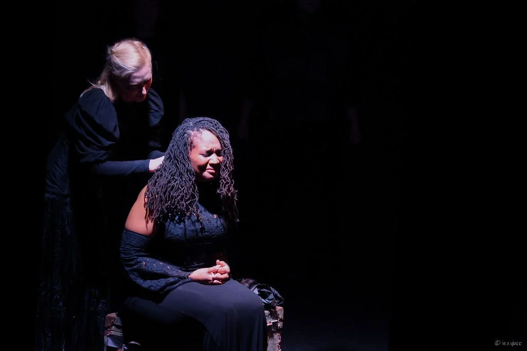 A woman with curly hair sitting on a chair with a look of distress, while another woman with blonde hair in a black outfit leans over her offering comfort or support, on a dark stage with minimal lighting.