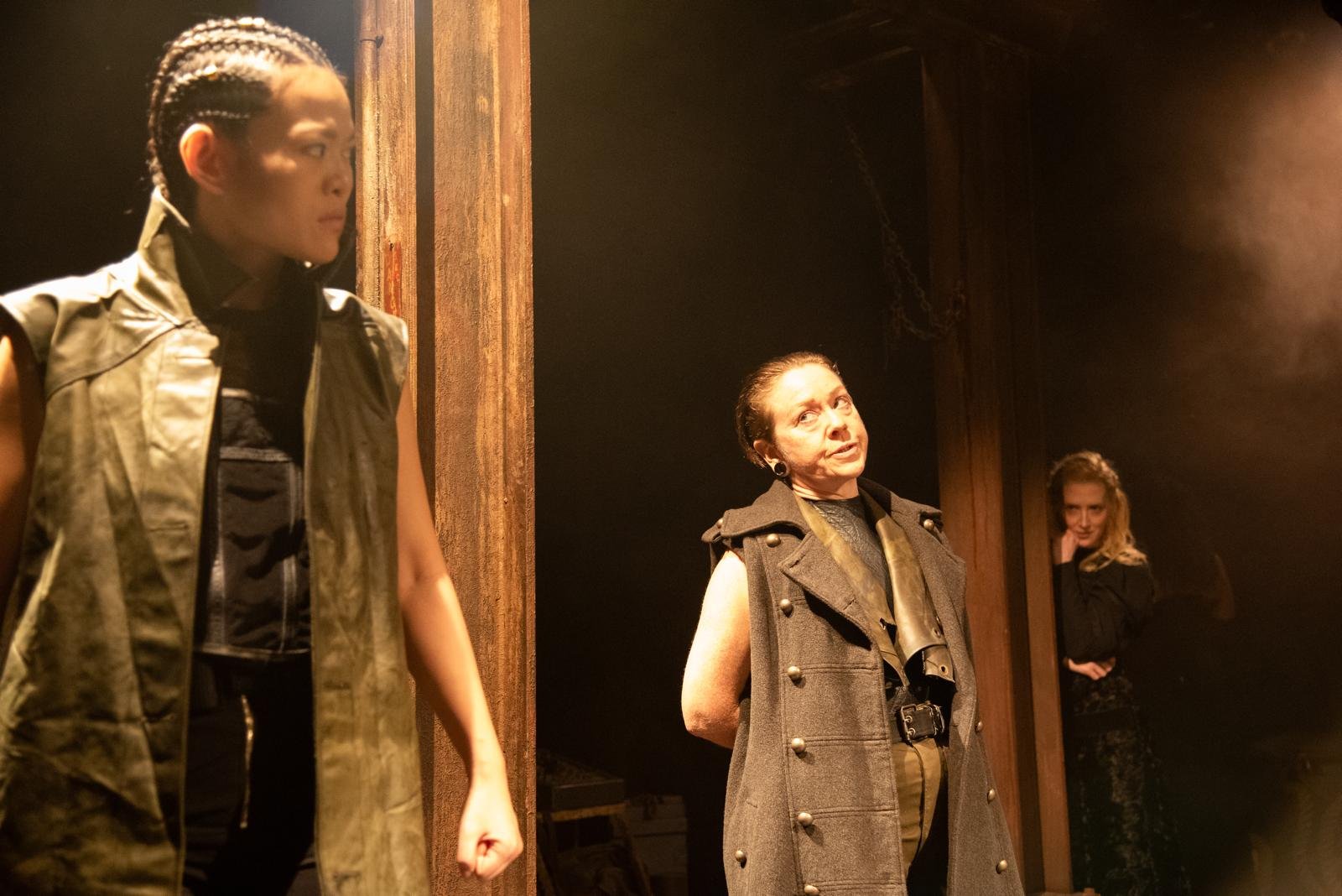Three women on stage, one young woman in military-style vest in foreground, middle-aged woman in gray coat with buttons in center, and another woman with blonde hair in black background.